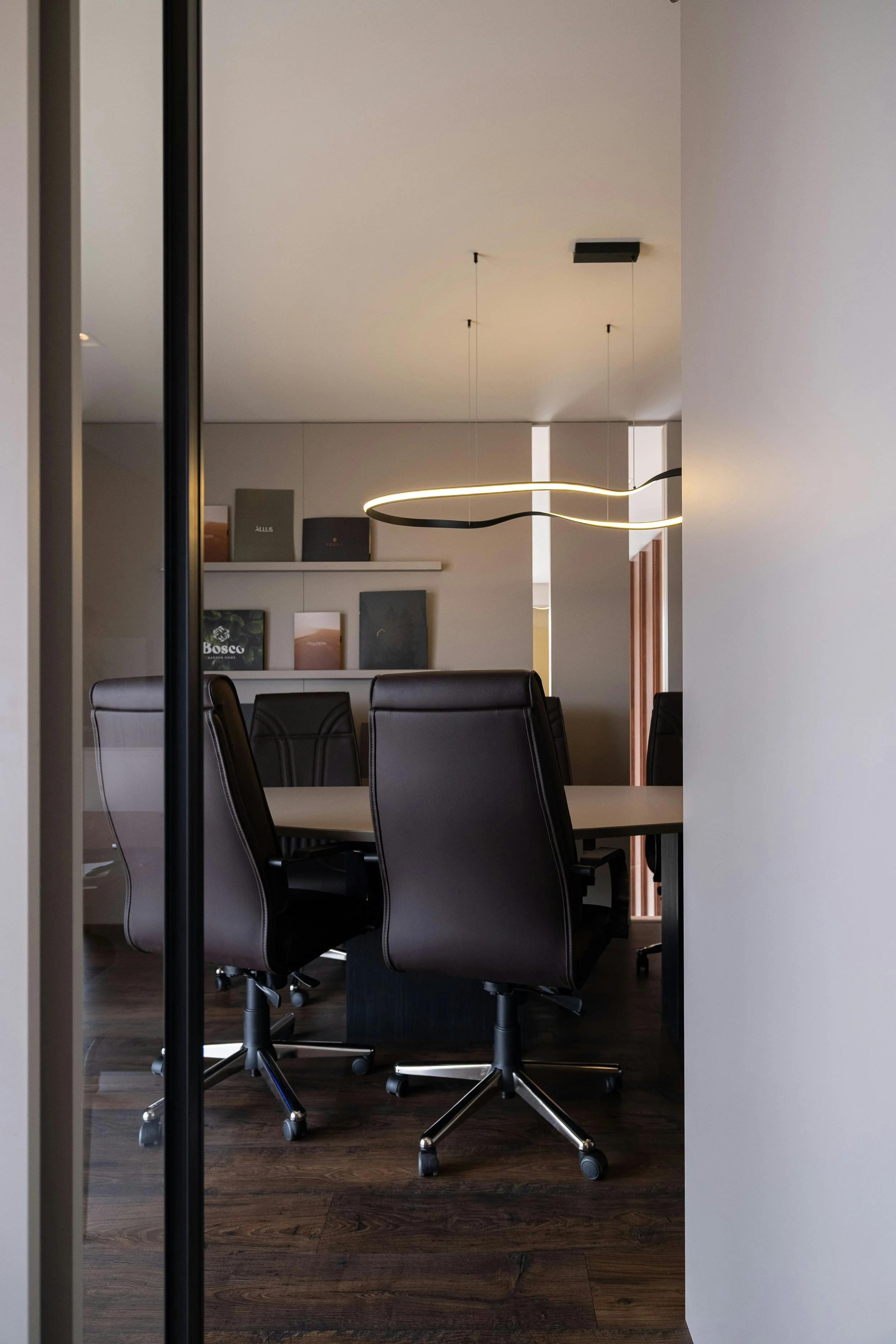 A modern conference room with a dark wooden table surrounded by black leather office chairs on casters. In the background, there are bookshelves with books and magazines, and a curved LED ceiling light fixture hanging from the ceiling.