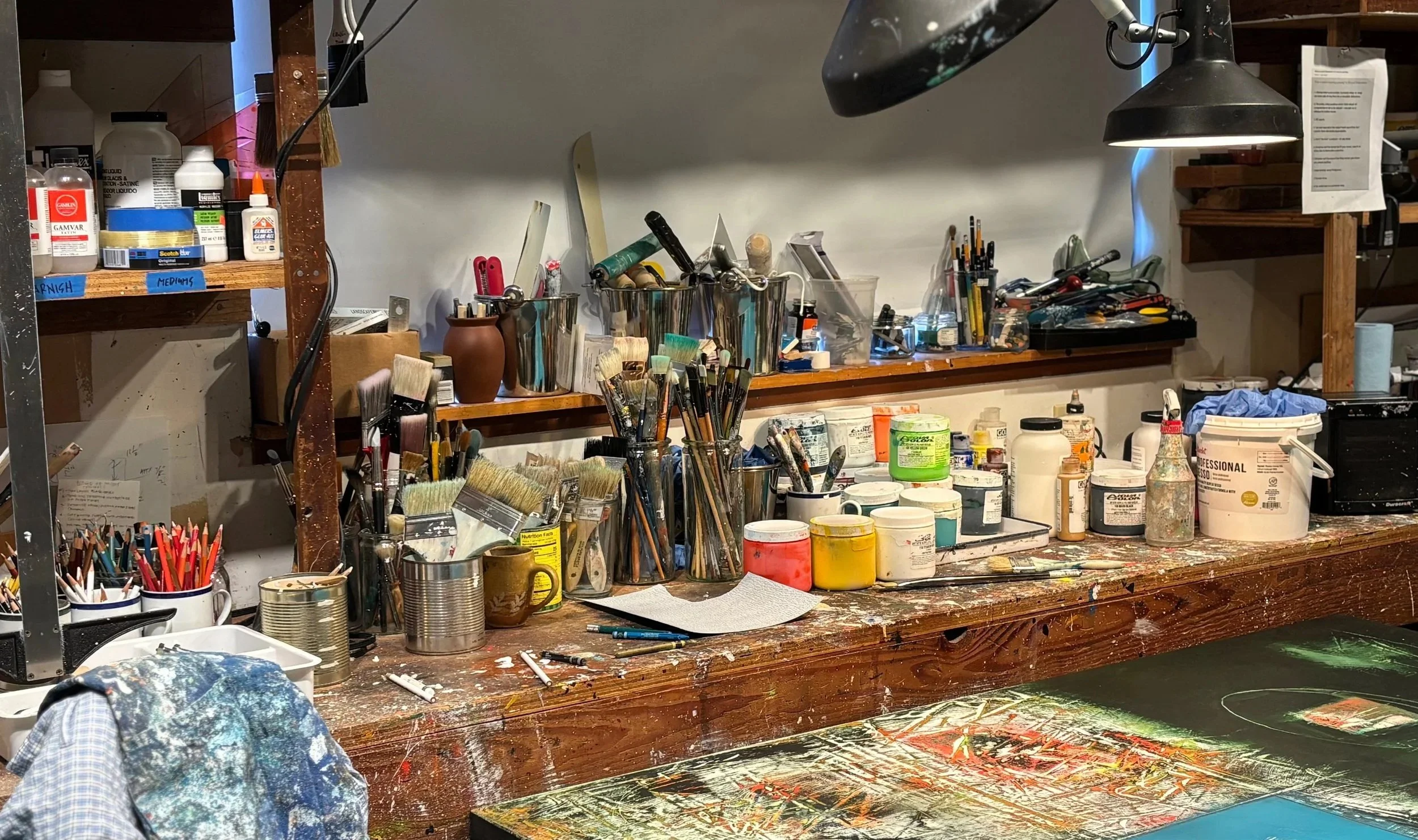 Art studio workspace with paintbrushes, tools, and supplies on a wooden table and shelf, with paint splatters and a partially finished artwork on the table.