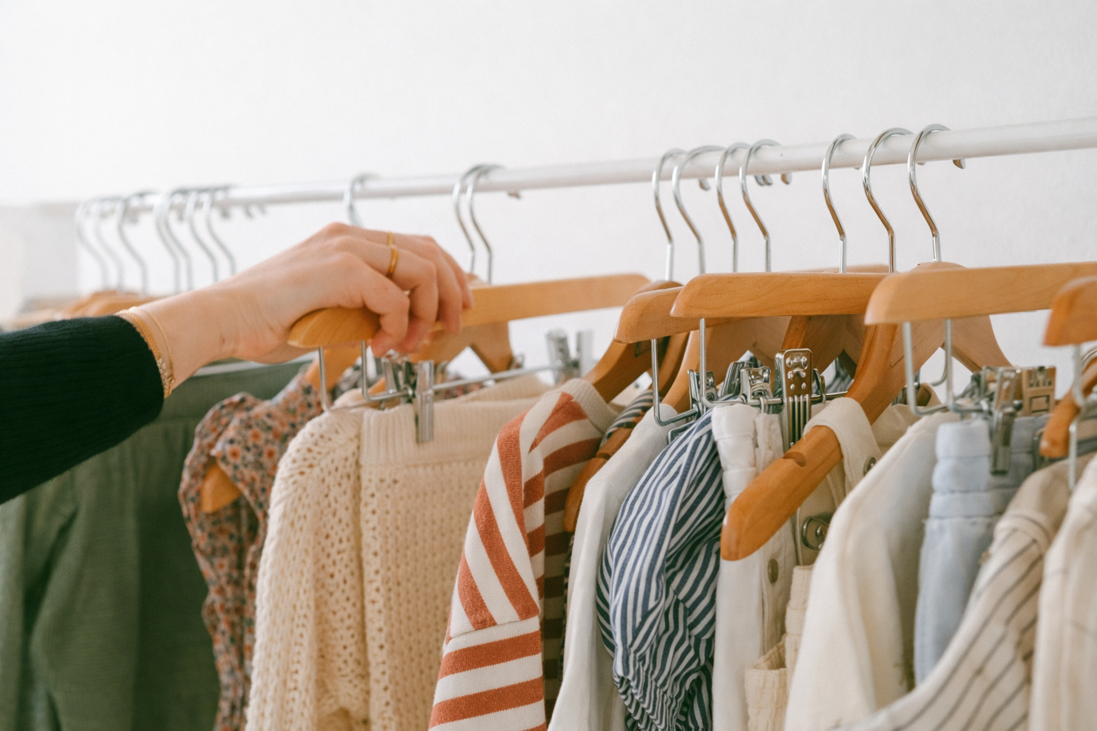 A person wearing a black sweater shops for clothes on a hung rack filled with casual shirts and sweaters, featuring various colors and patterns.