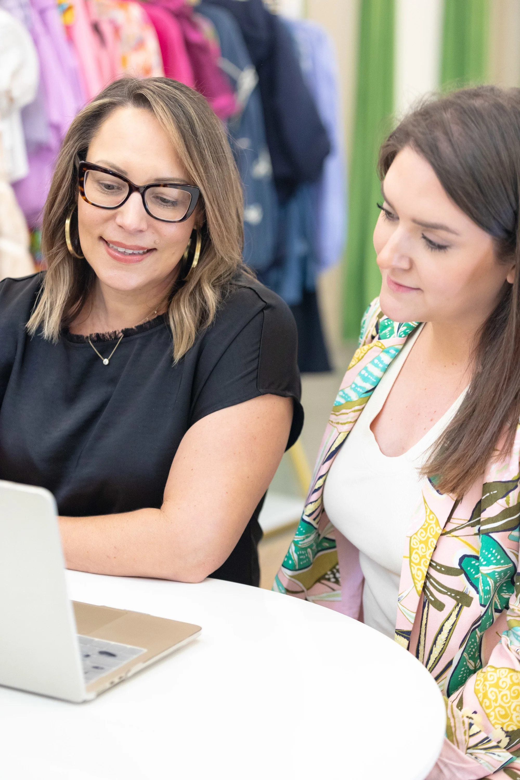 Two women looking at a laptop together at a table in a store with clothing racks in the background.