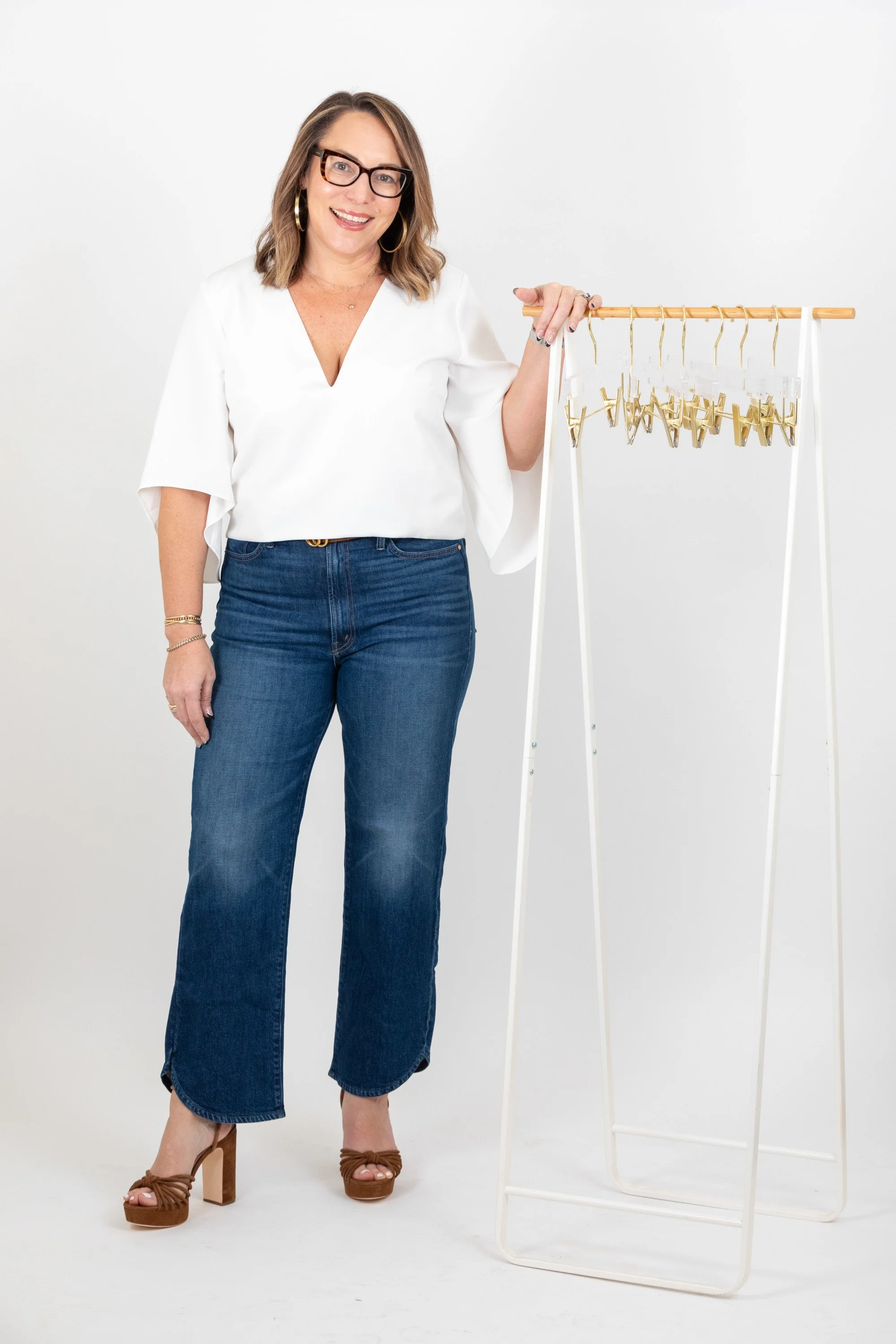 A woman with glasses, wearing a white top and jeans, standing next to a clothing rack with gold hangers.