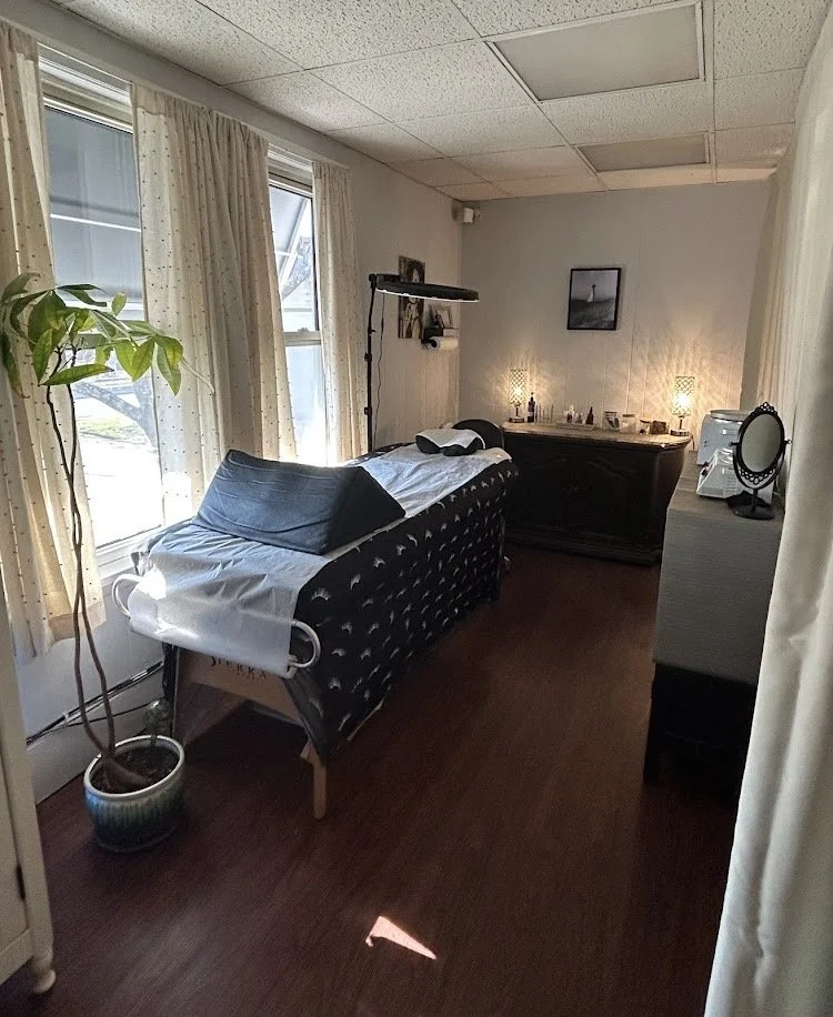 A room with a massage table near window, side table with mirror, and a dresser with lamps and decorative items.