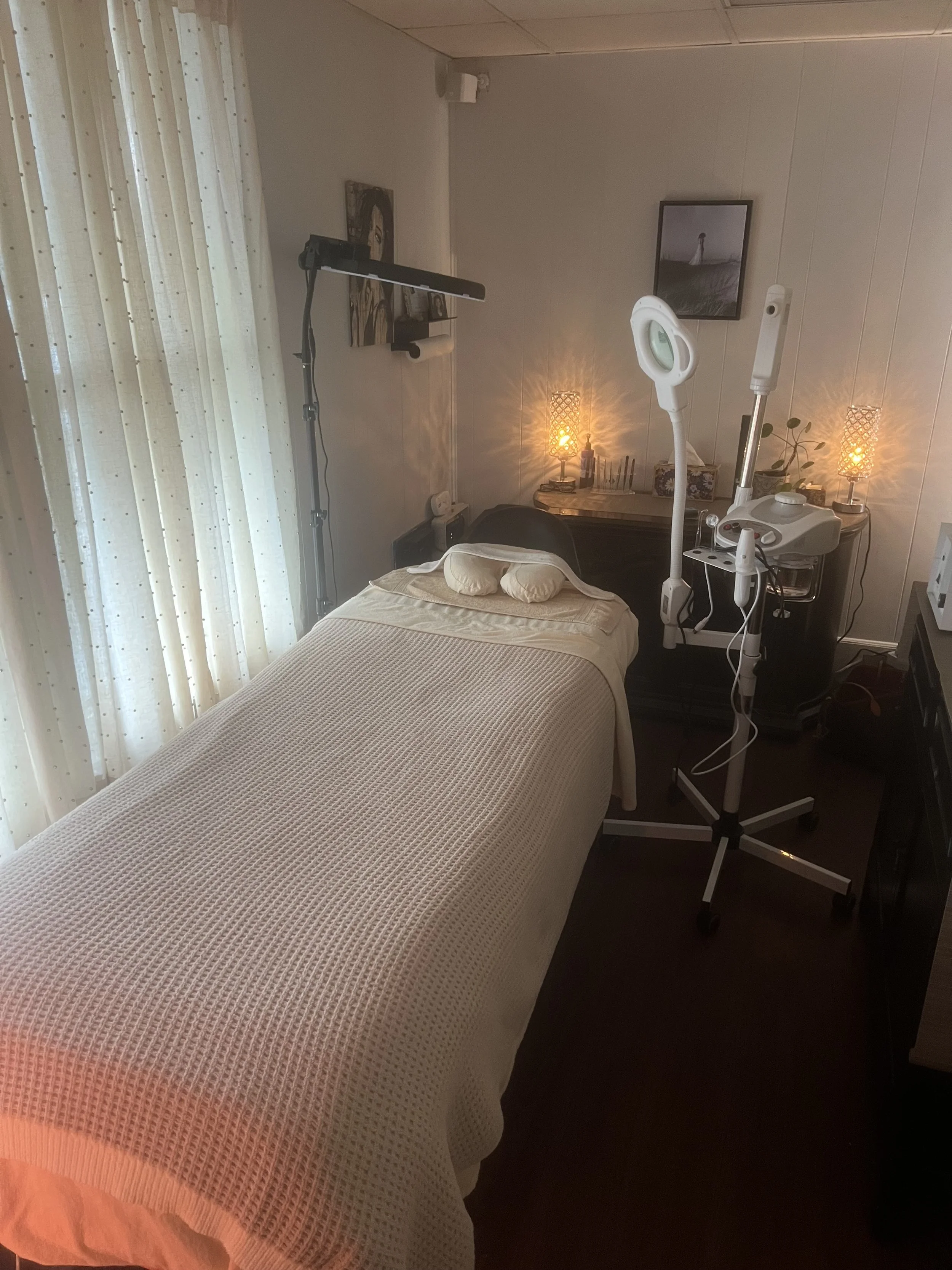 A massage therapy room with a massage table covered in a cream-colored blanket, a headrest at the pillows, and soft lighting from lamps and candles. Equipment and artwork are visible on the walls and surface near the table.