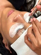 A person receiving a facial treatment with cotton pads on their face, while a professional applies a skincare procedure with a brush.