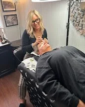 A woman with blonde hair and glasses is giving a manicure to a person lying on a reclined chair in a salon. The woman is smiling and appears to be beautifying the person's hands.