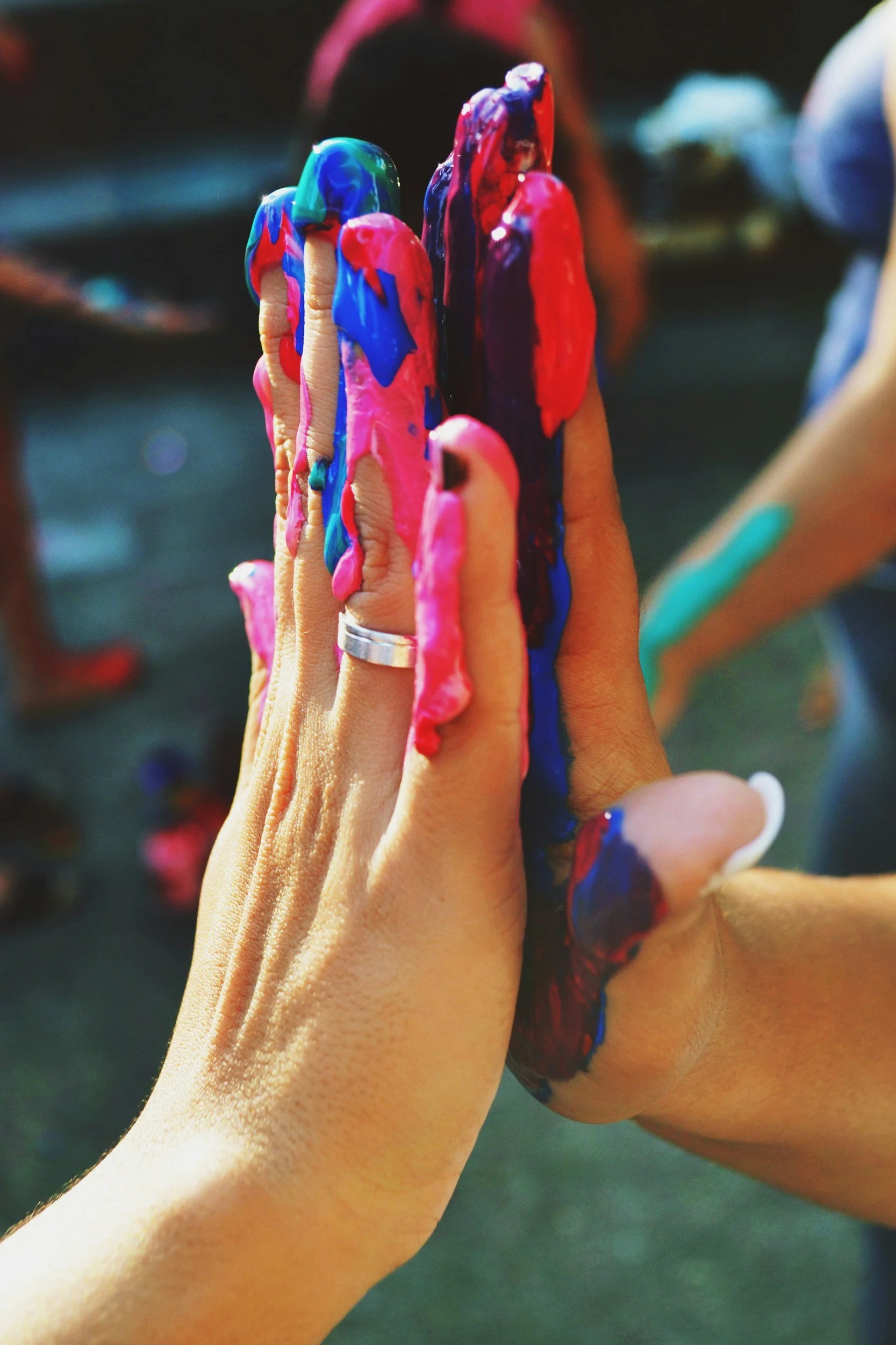 Two hands pressed together in a high five with colorful, smeared paint on fingers and palms, in an outdoor setting with blurred background.