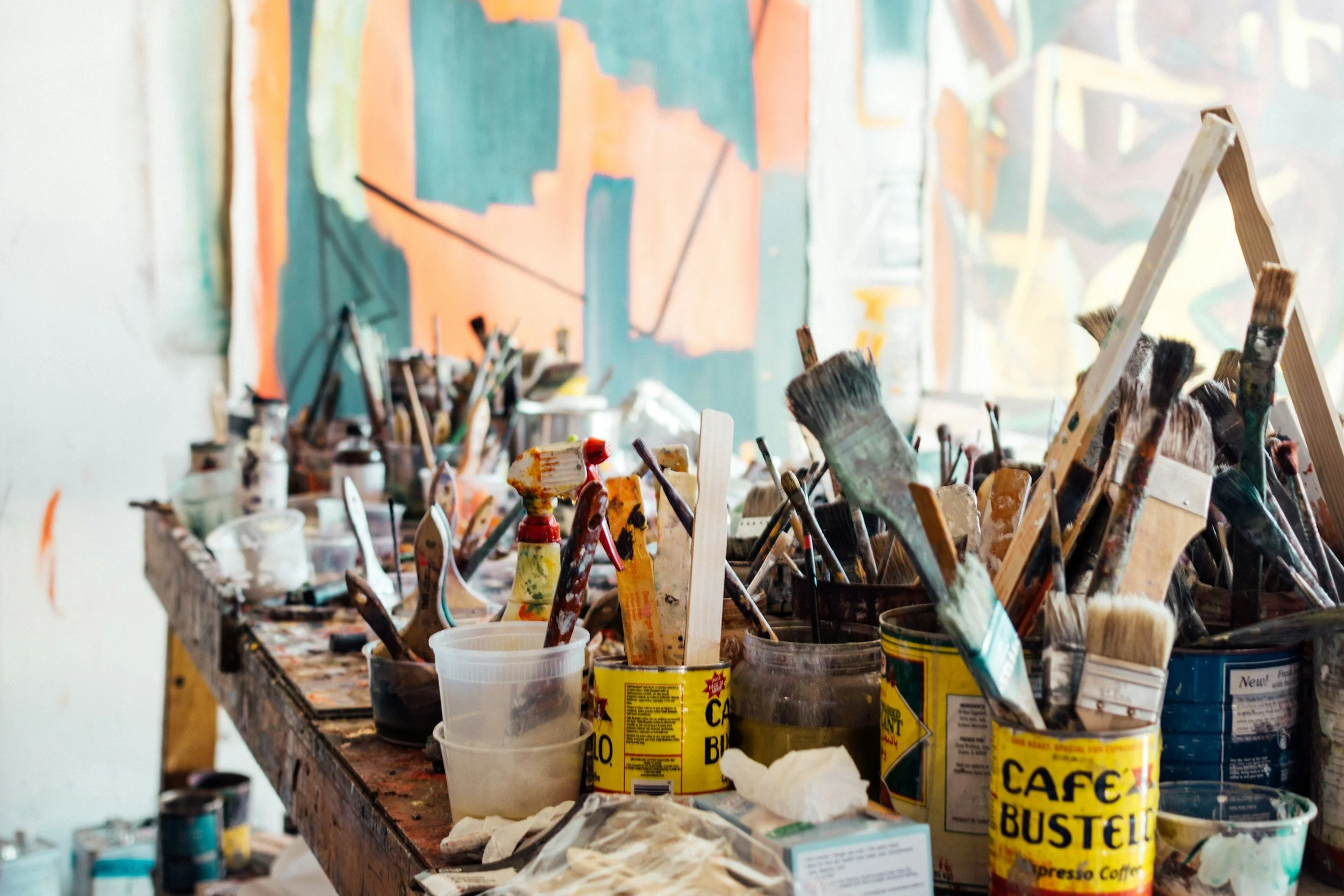 Art supplies scattered on a cluttered work surface, including paint brushes, mixing containers, and art jars, with a colorful abstract painting in the background.