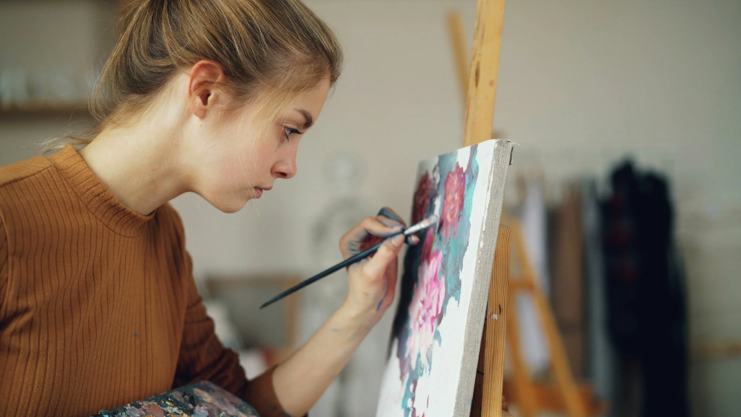 A woman with blonde hair tied back, wearing a brown long-sleeve shirt, is focused on painting a colorful canvas with a small brush in an art studio.