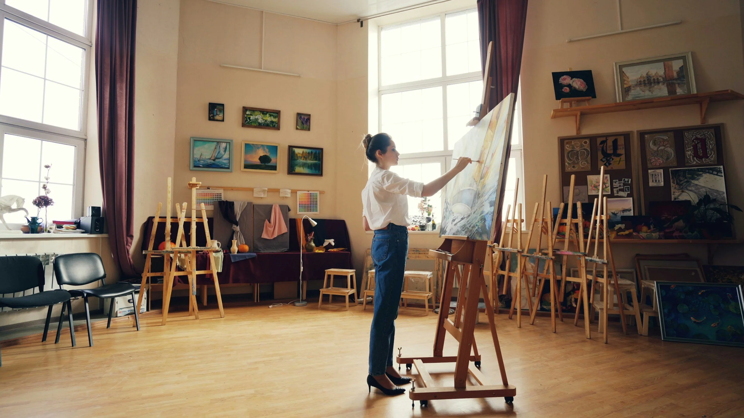 A woman painting on a large canvas in an art studio filled with paintings, easels, and art supplies.