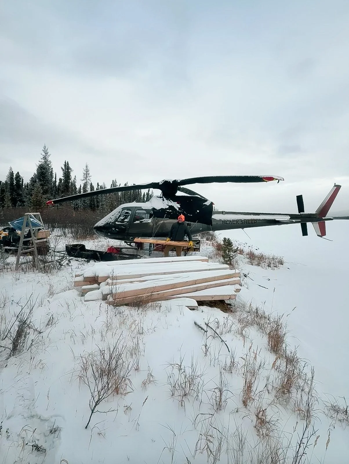 A helicopter on snowy ground with logs stacked nearby, a person in safety gear, and a forest background under an overcast sky.