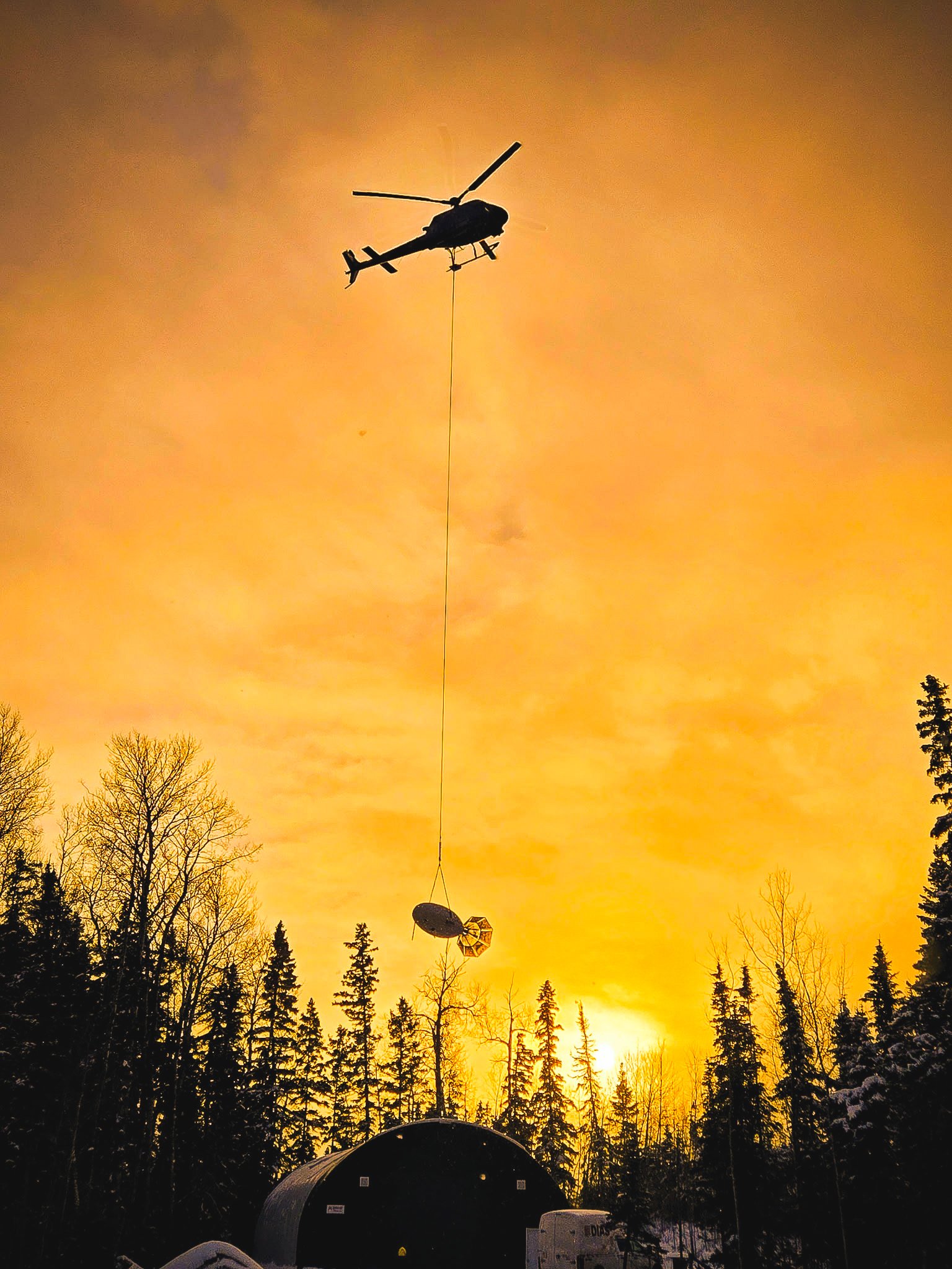 Helicopter flying in the sky at sunset, carrying a large satellite or dish antenna.