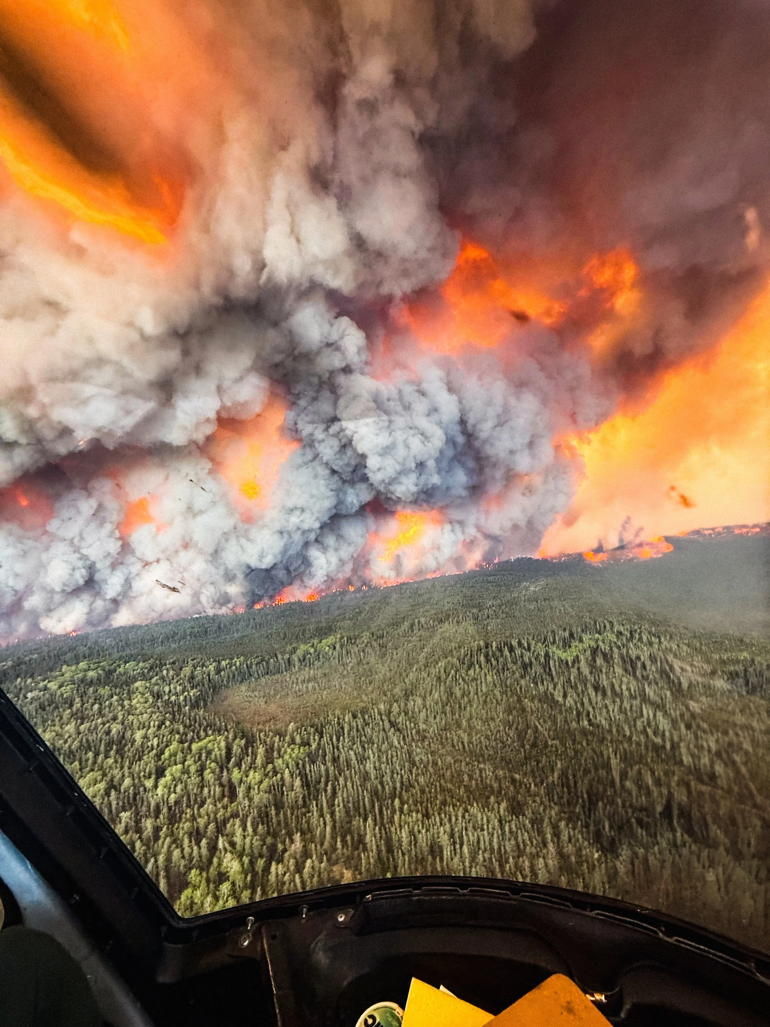 View from an aircraft cockpit showing a wildfire with thick smoke and flames over a forested mountain.