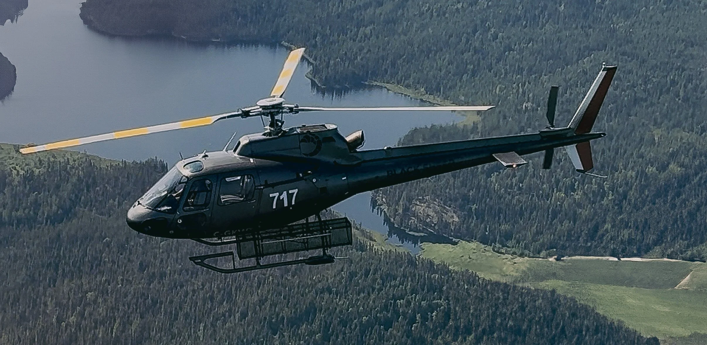 A black helicopter flying over a landscape with a lake and forested area.