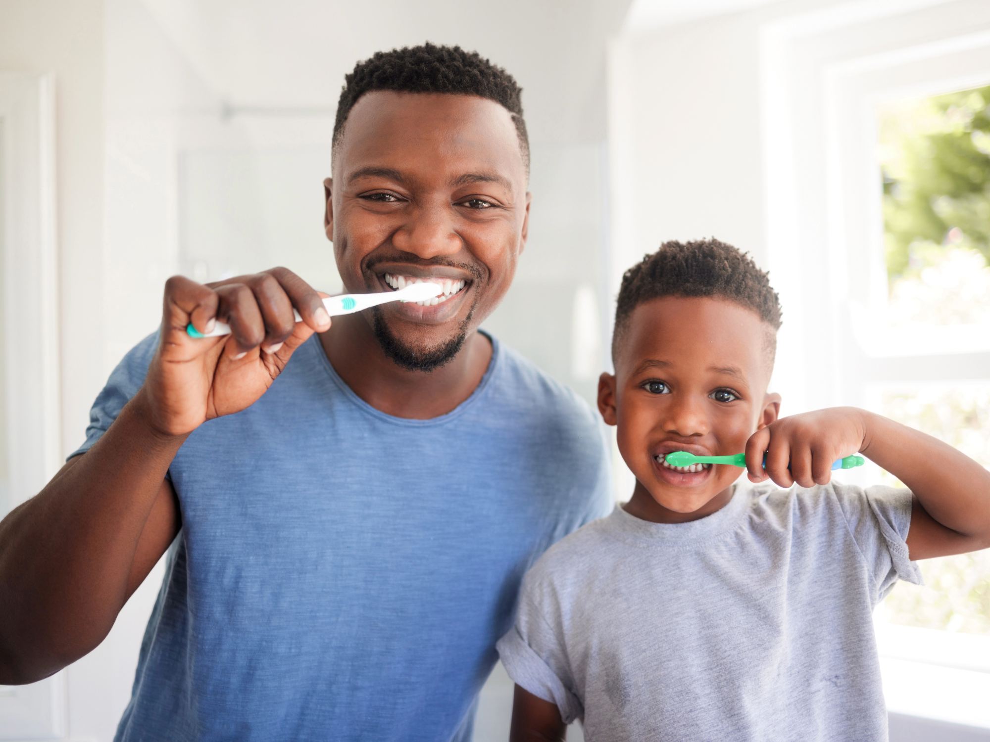 A smiling man and a smiling boy standing together in a bright bathroom, brushing their teeth with toothbrushes.