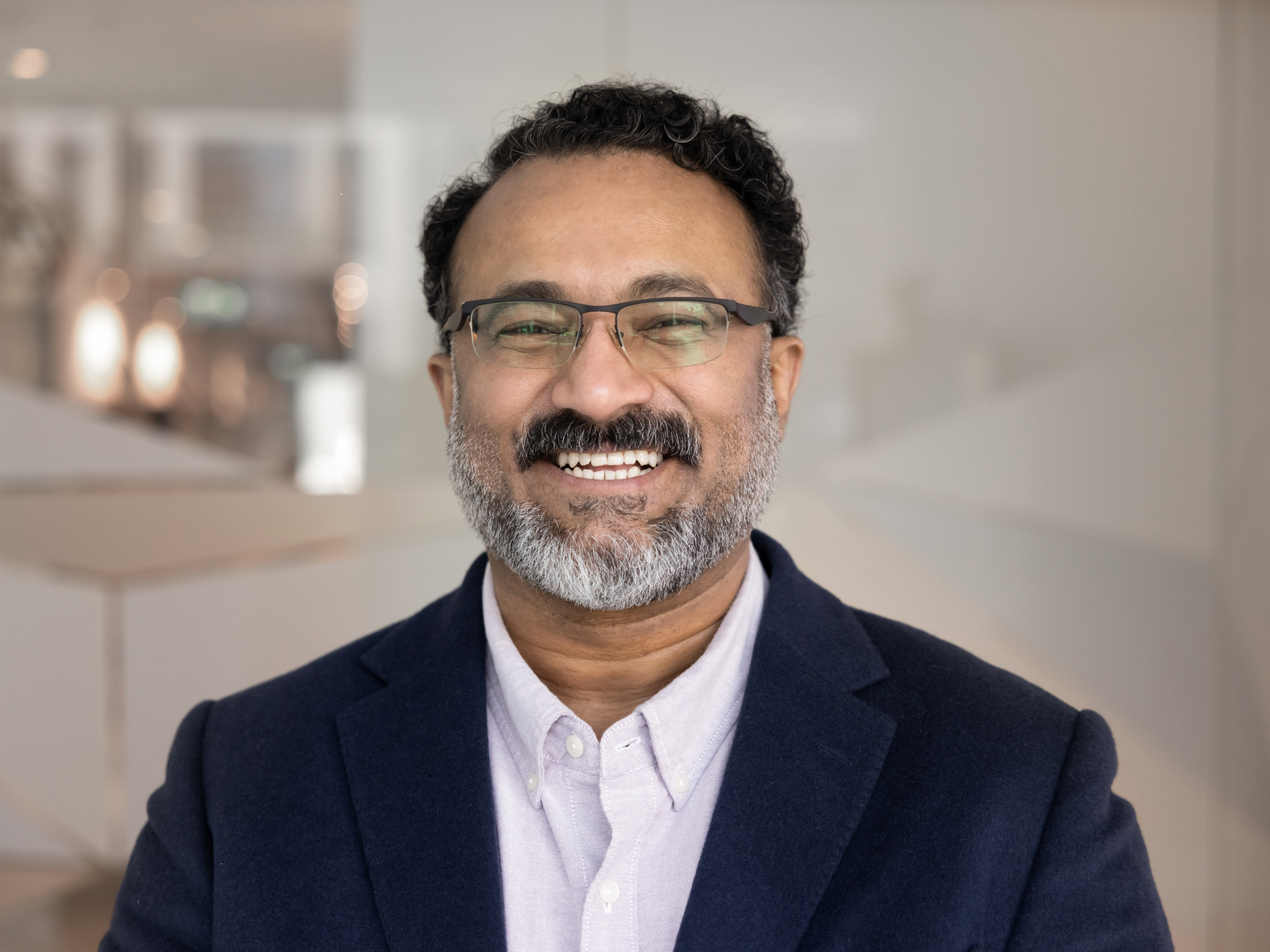 A smiling middle-aged man with glasses, a beard, and dark curly hair, wearing a navy blazer and a white collared shirt, in a bright indoor setting.