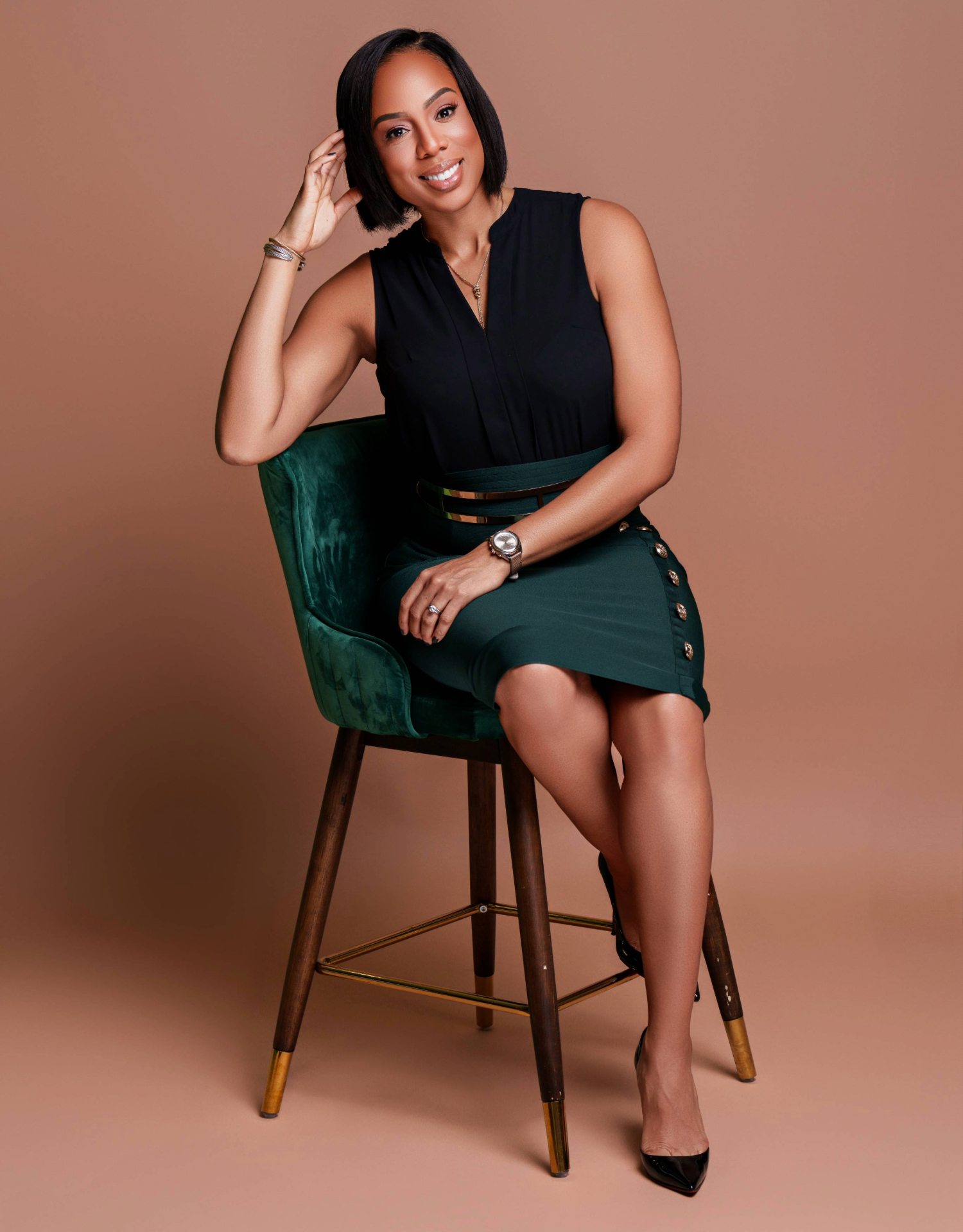 A woman sitting on a green velvet chair with a beige background, wearing a black sleeveless top and green skirt with buttons, smiling and looking at the camera.