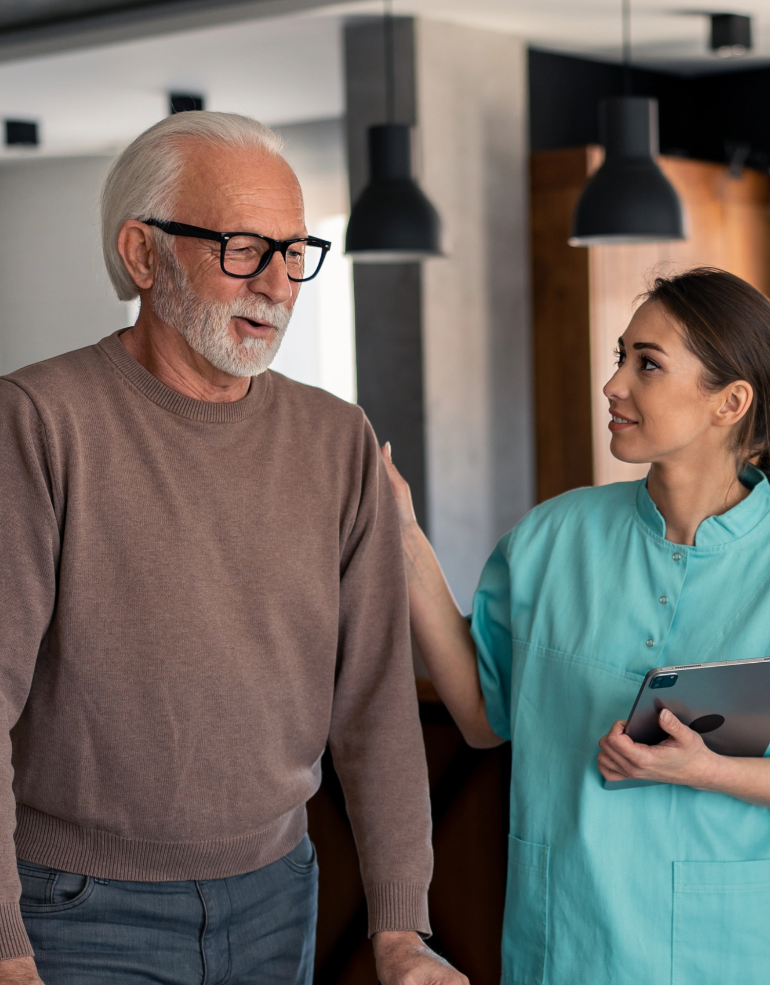A healthcare professional, a young woman, gently places a hand on an elderly man's shoulder while holding a tablet, and they share a warm moment together in a modern indoor setting.