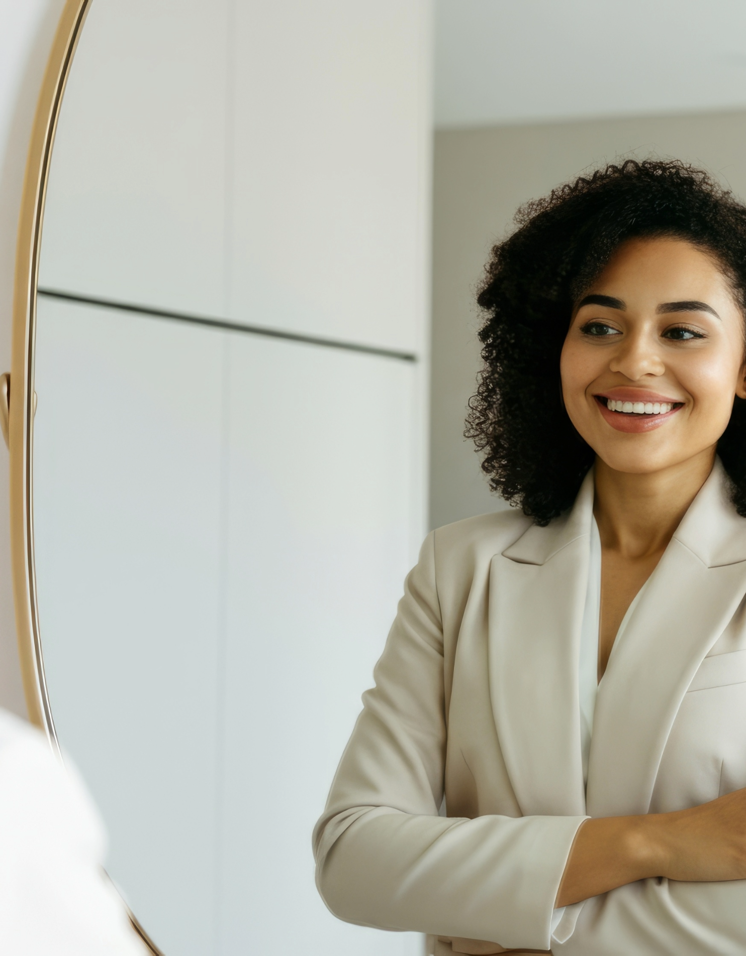 A woman looking at herself in a mirror with a smile, wearing a beige blazer in a modern room.