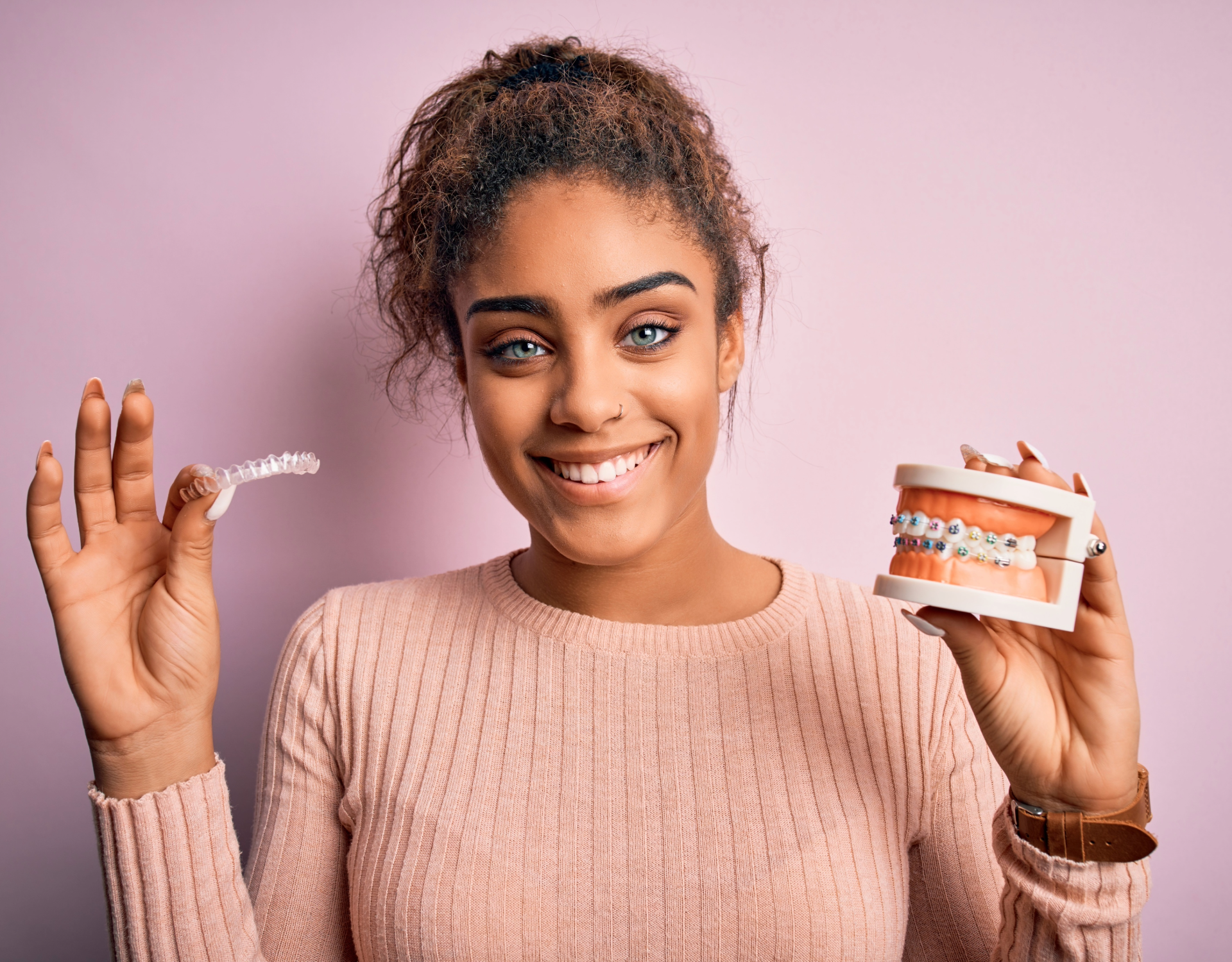 Young woman smiling and holding a dental retainer in one hand and a model of teeth with braces in the other, against a pink background.