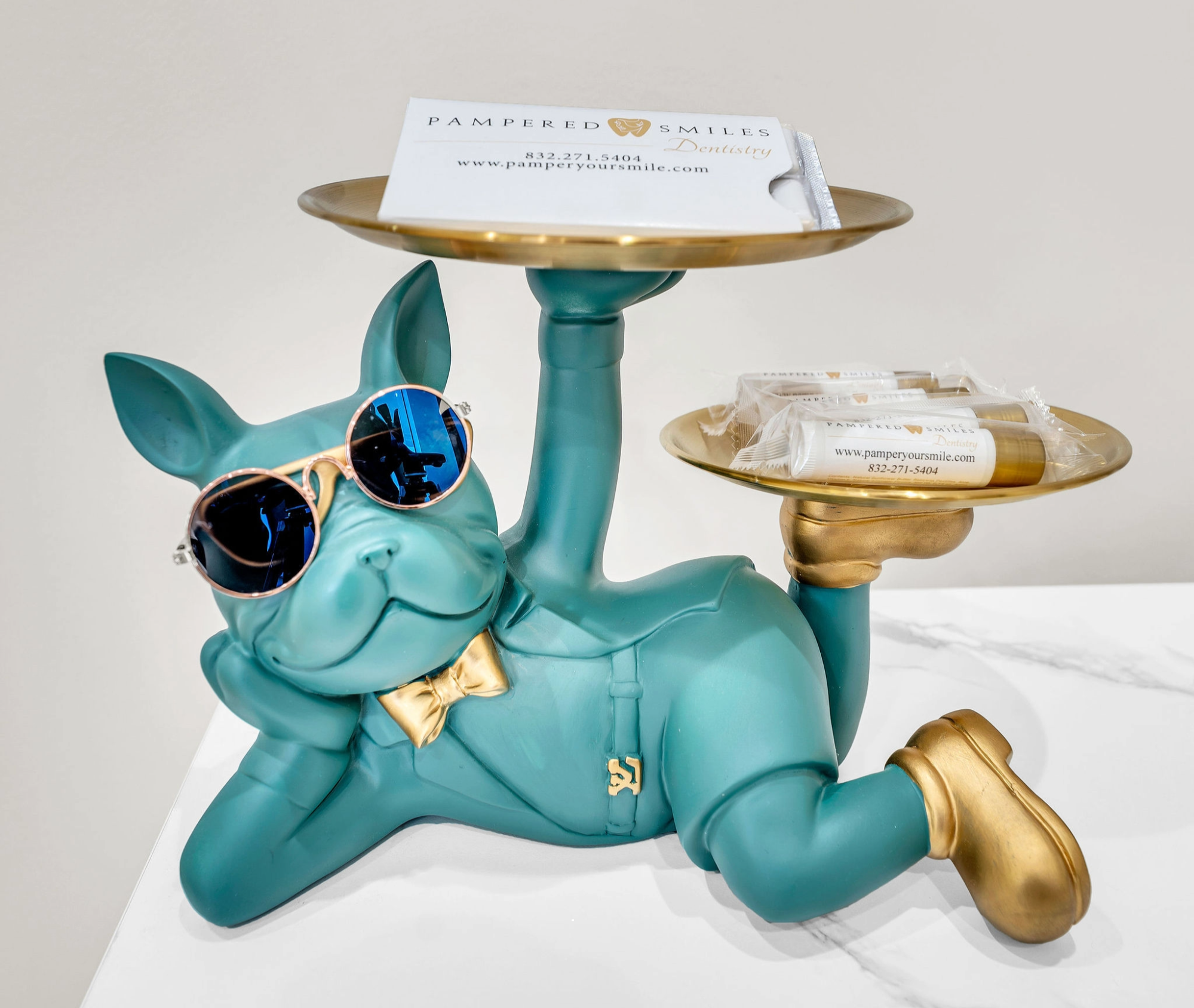 A teal-colored bulldog figurine with sunglasses and a gold bow tie, lying on its side with one arm supporting its head, dressed in a gold vest, gold shoes, and gold accents. It holds two gold trays, one on its raised paw with business cards and the other with small packaged items.