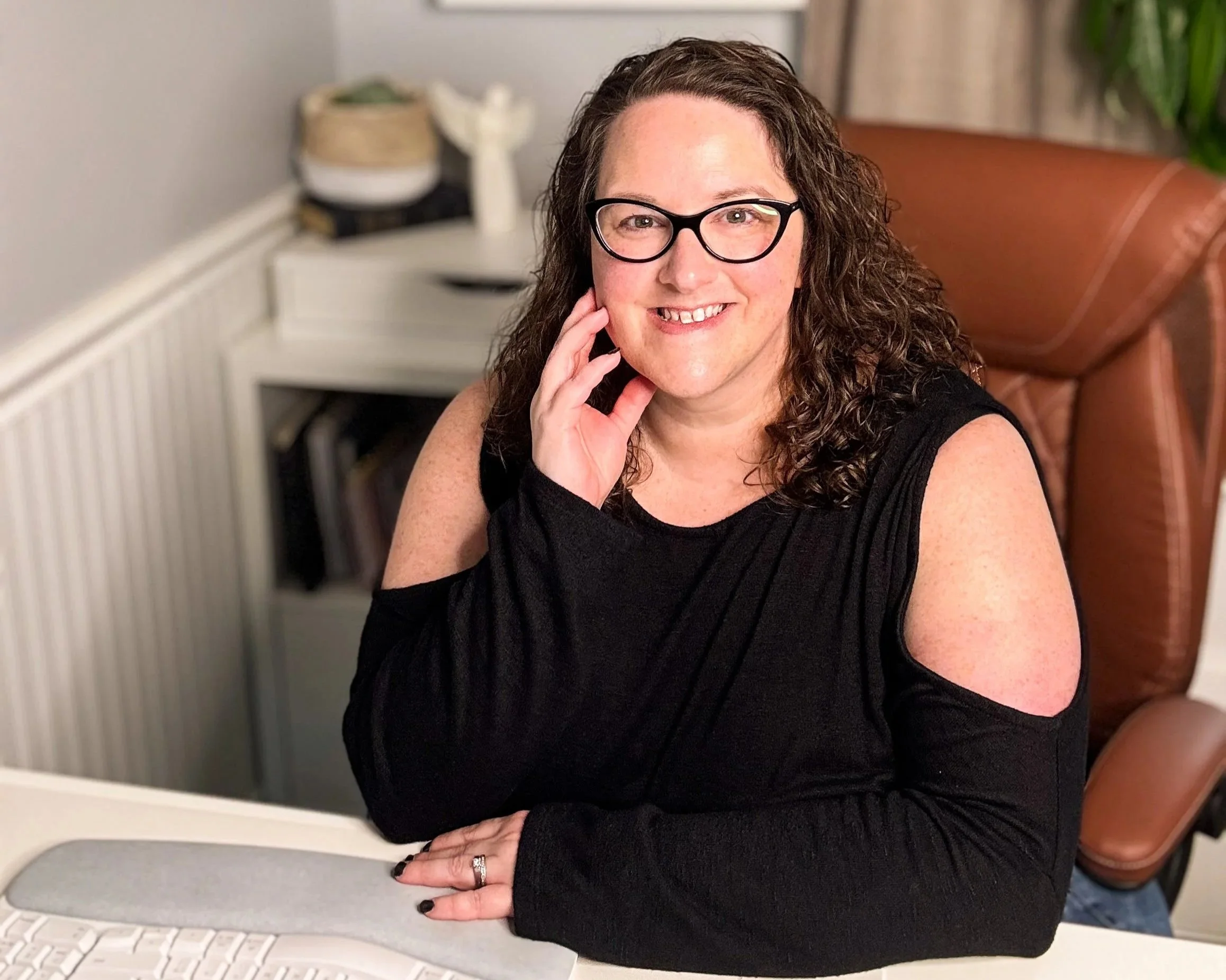 A woman with curly brown hair, wearing glasses and a black cold-shoulder top, sitting at a desk and smiling at the camera.