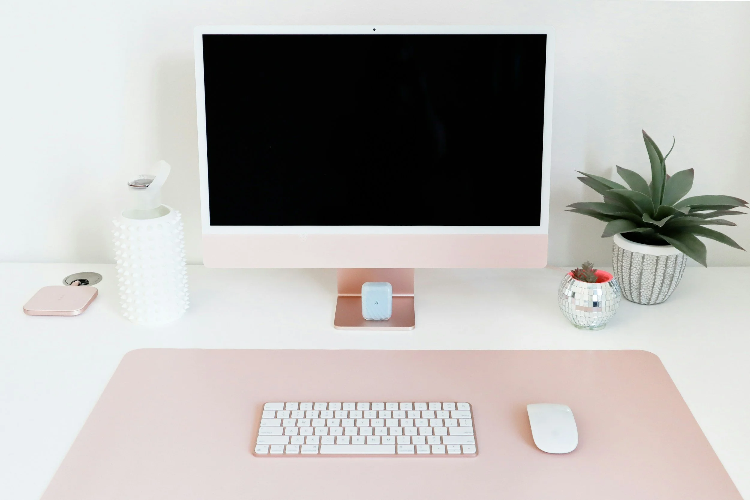 A workspace with a pink and white color scheme, featuring a computer monitor, wireless keyboard and mouse, decorative plants, a small disco ball, a white textured bottle, and a pink coaster with a cup on it.