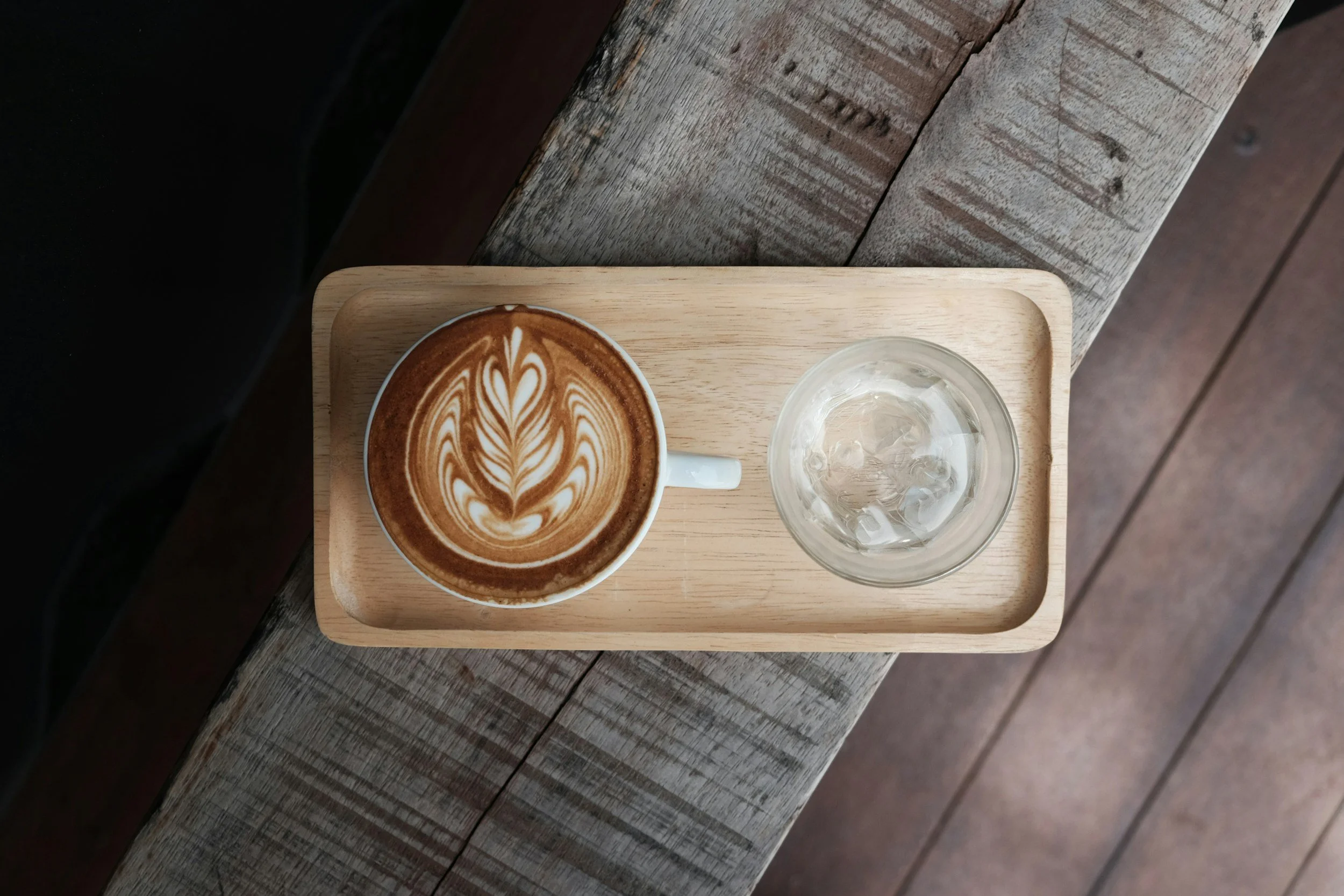 A latte with latte art and a glass of water on a wooden tray, placed on a rustic wooden table.