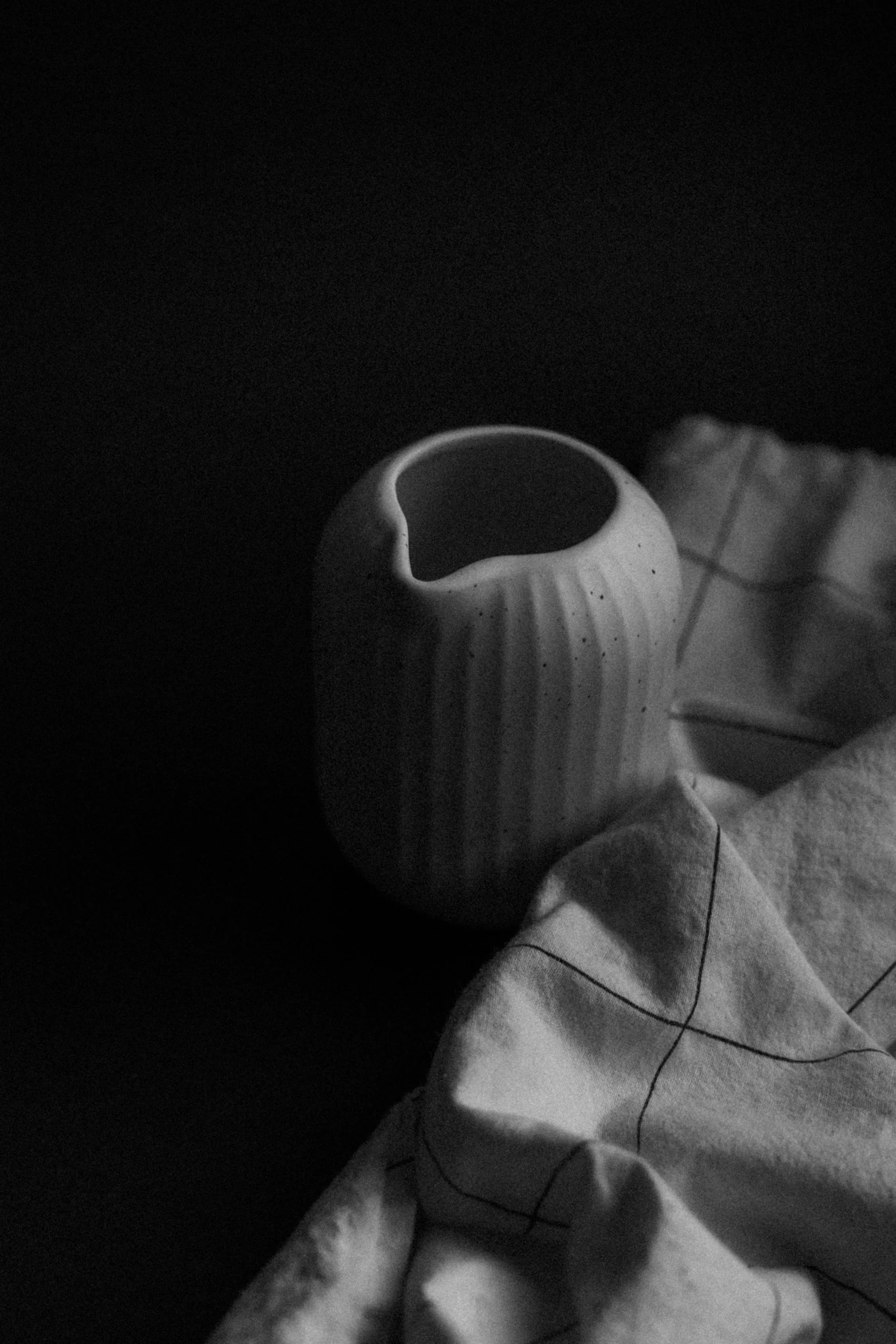 Black and white photo of a ceramic vase with vertical ridges and a broken rim, accompanied by a piece of cloth with grid lines.