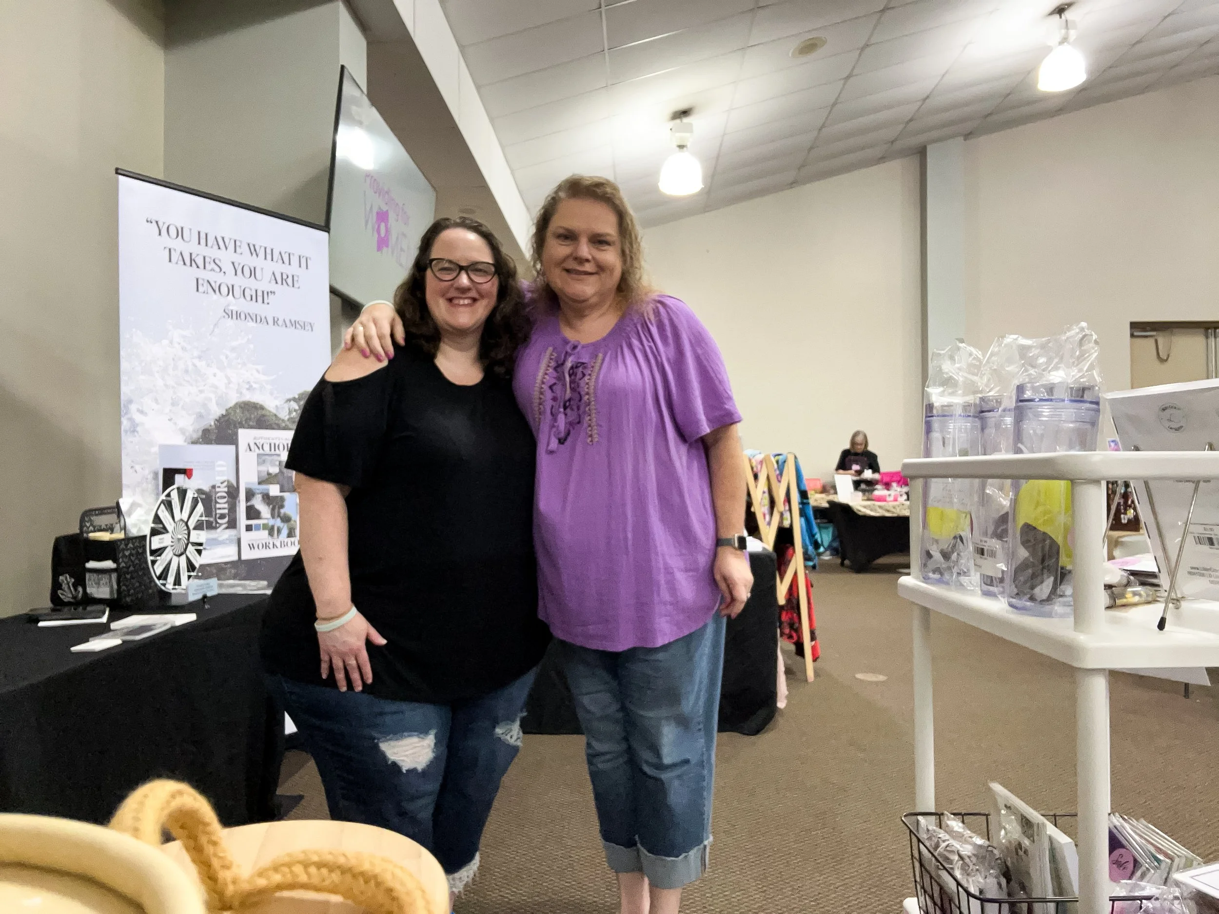 Two women smiling and standing close together indoors at an event, with tables and displays in the background.