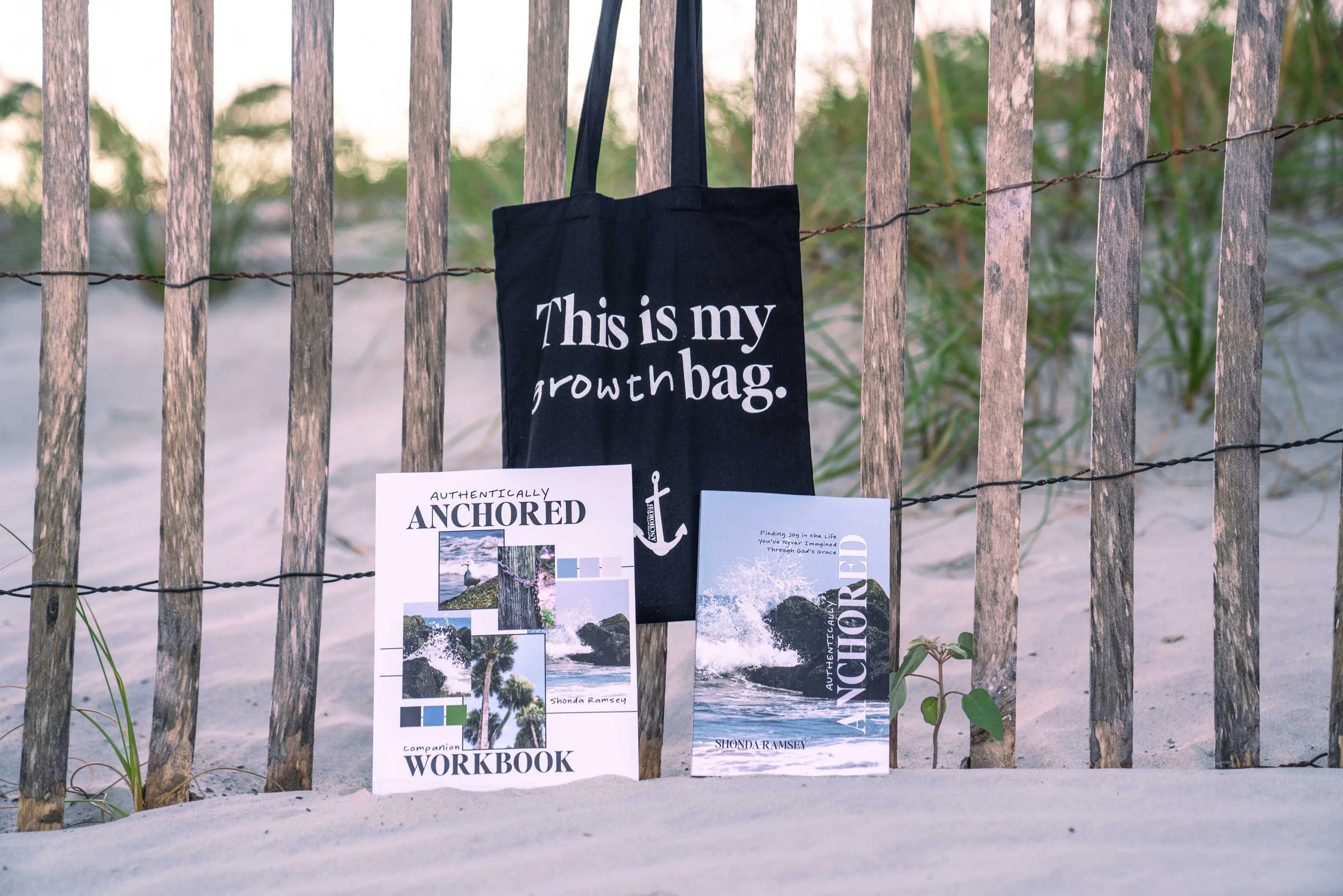 A black tote bag with white text that says 'This is my growth bag' hanging on a wooden fence near a sandy beach with green plants in the background. Two books titled 'Authentically Anchored' and 'Authentically Anchored' by Shonda Ramsey are placed on the sand.