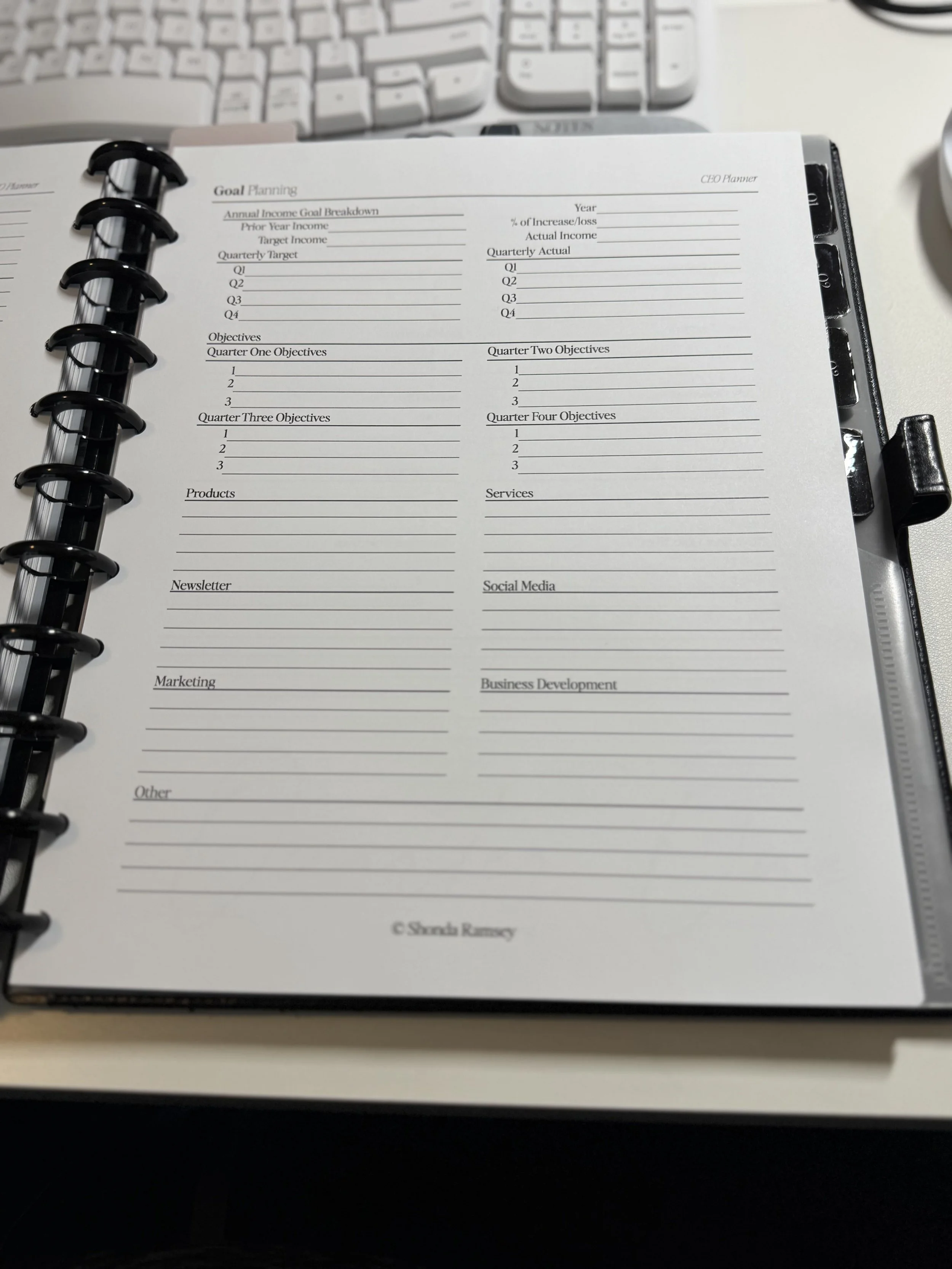 A printed goal planning sheet titled 'Goal Planning' with sections for annual income goals, quarterly targets, objectives, products, newsletter, marketing, other, services, social media, and business development, situated on a desk next to a white keyboard.