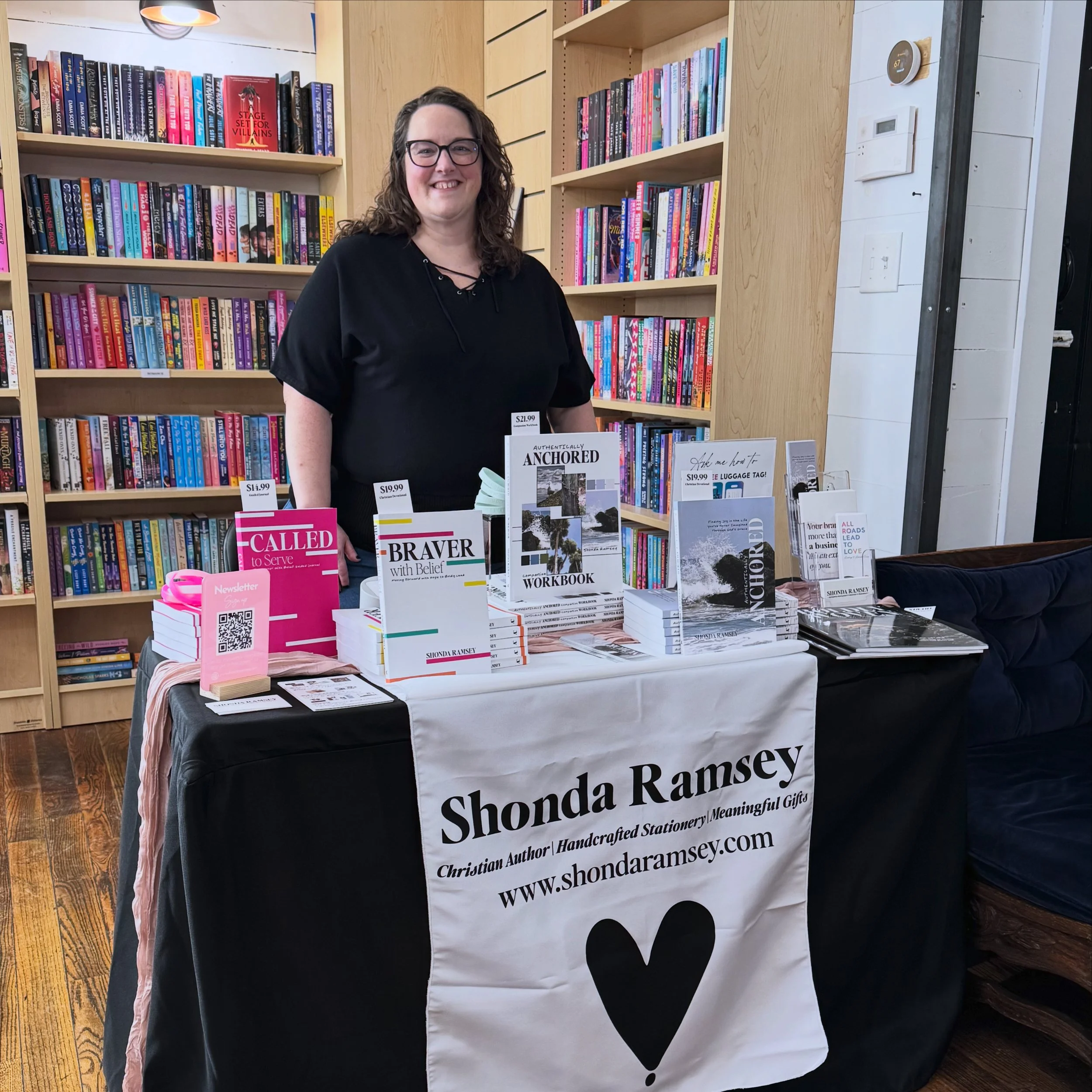 A woman with glasses and curly hair standing behind a table with books and signs at a book signing event. Books and signs are displayed on the table, with a bookshelf filled with colorful books in the background. The table has a black tablecloth with white text that includes her name, Shonda Ramsey, and her website.