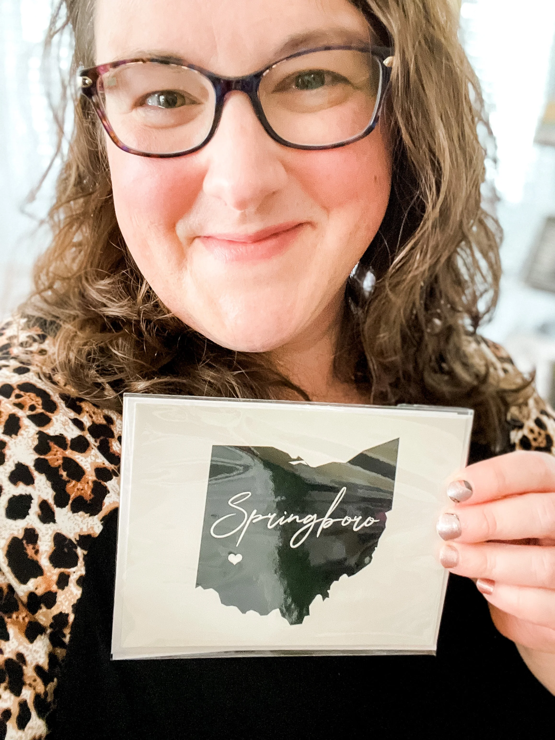 A woman with curly hair and glasses holding a sign with a heart and the outline of Ohio, with the word "Springboro" written across it.