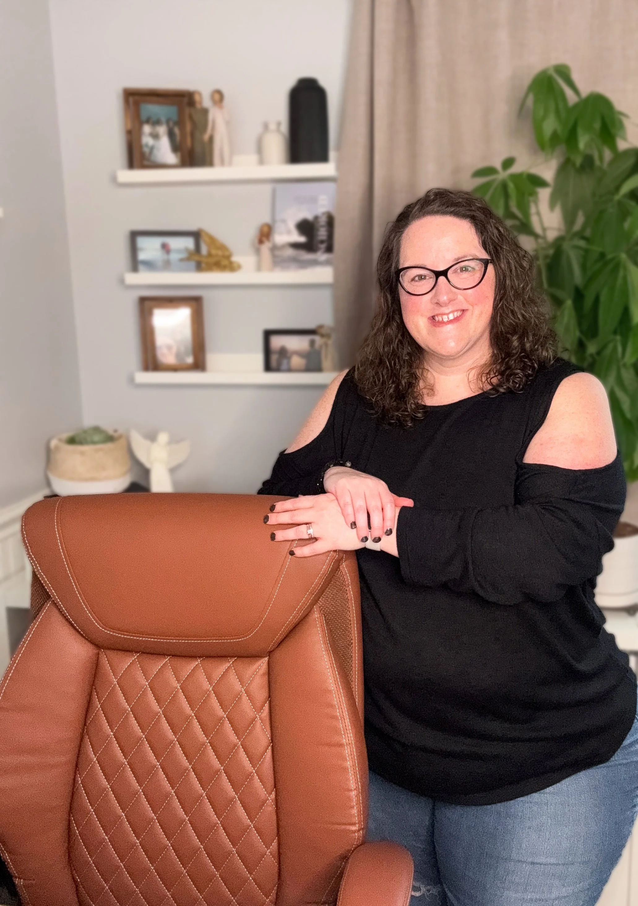 A woman with curly hair and glasses is smiling while leaning on a brown leather office chair in a room with shelves, framed photos, and a large green plant in the background.
