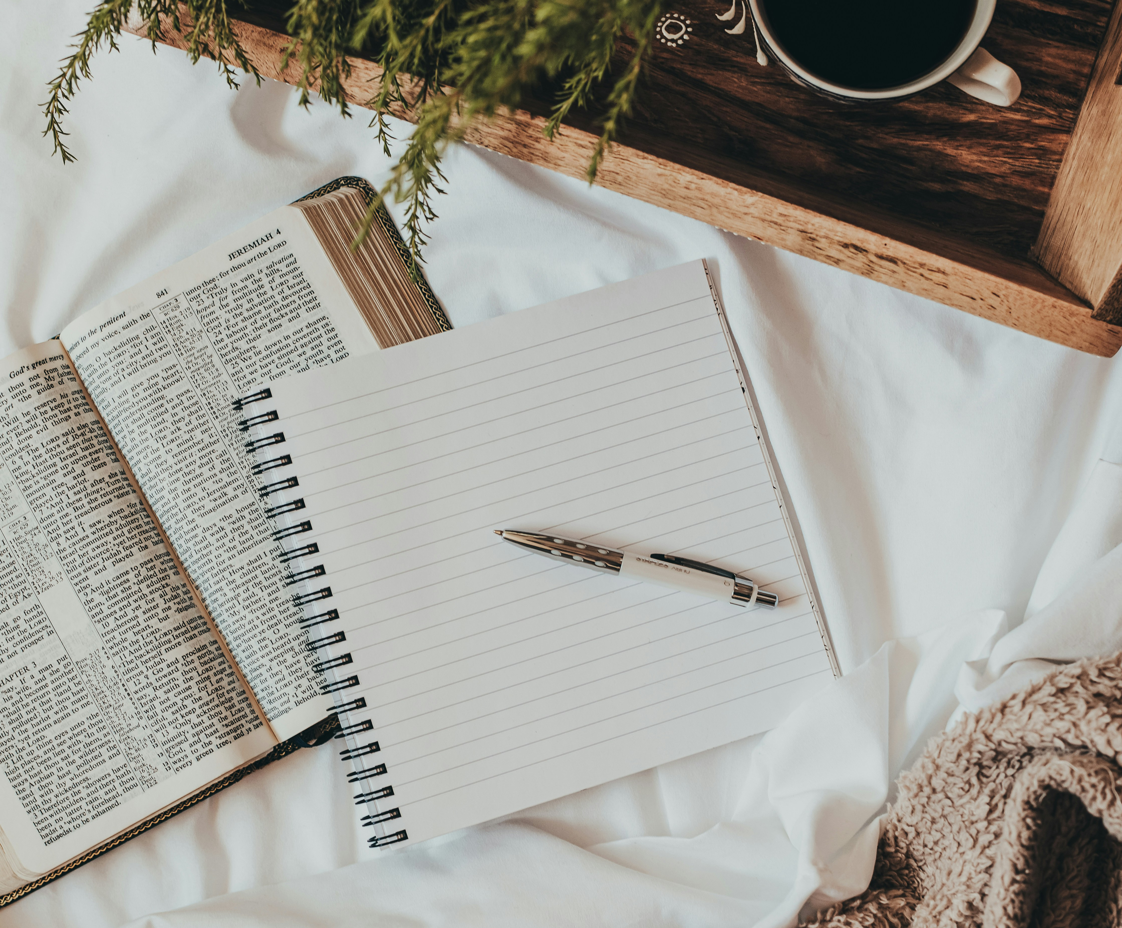 Open Bible, blank notebook with pen, and a cup of coffee on a wooden tray, with a blanket and some greenery.