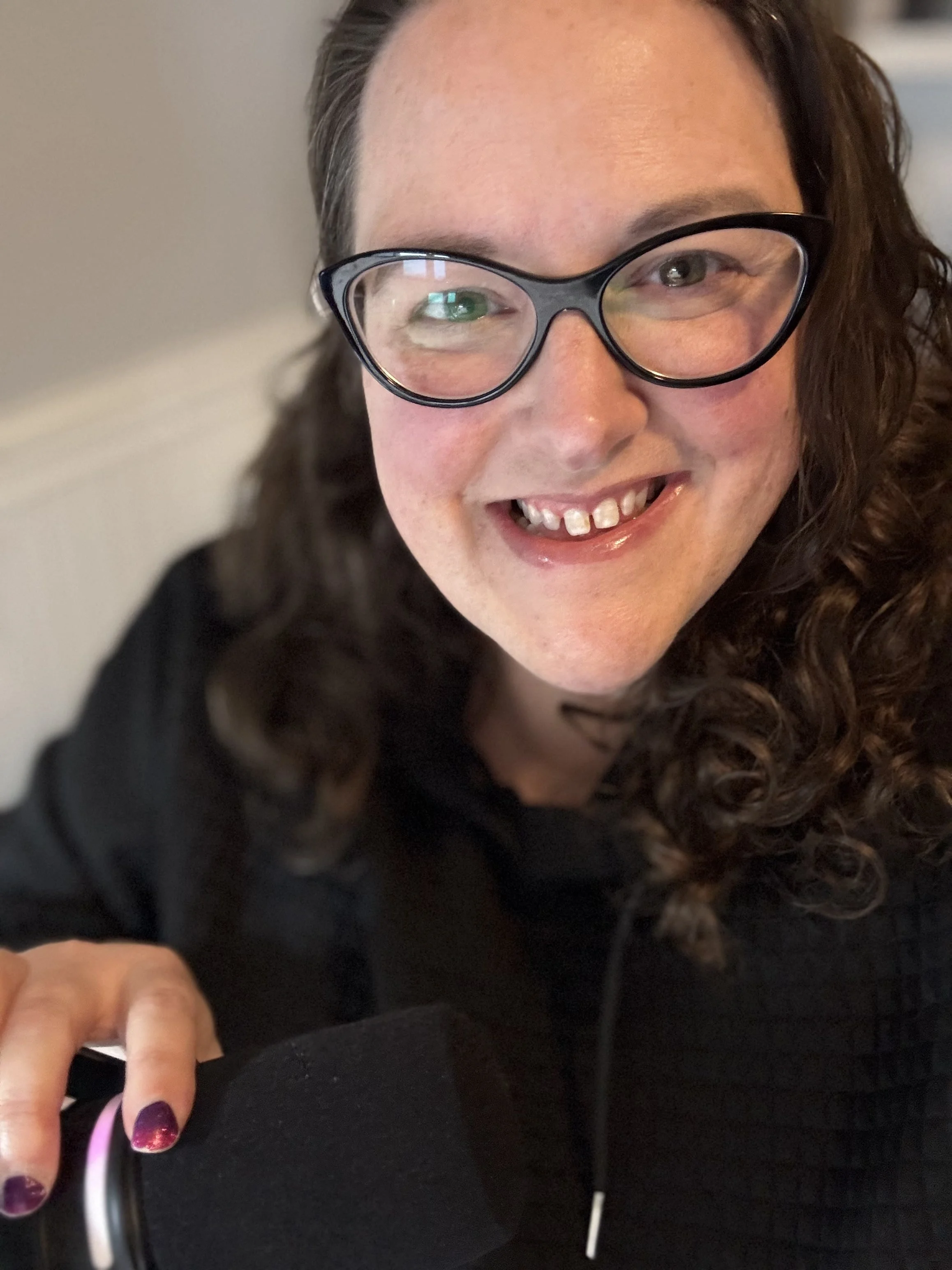 Close-up of a smiling woman with curly dark hair, wearing glasses with black frames, showing her teeth, and wearing a black top.