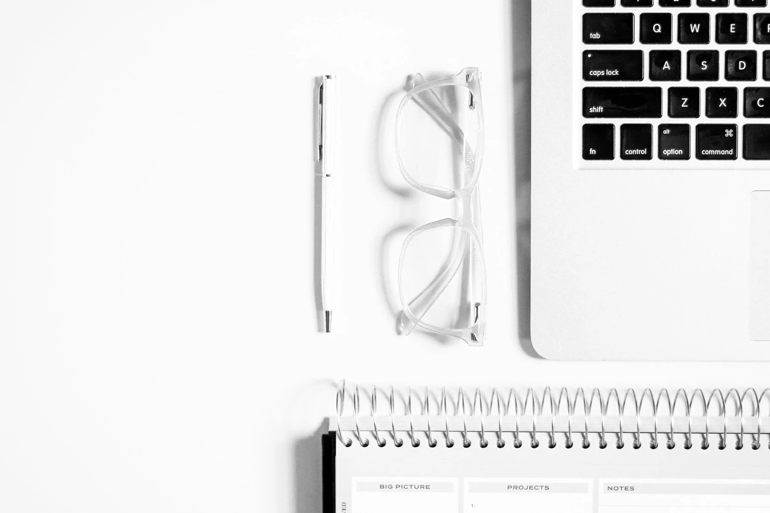 Desk workspace with a pen, eyeglasses, laptop, and spiral notebook on a white surface.