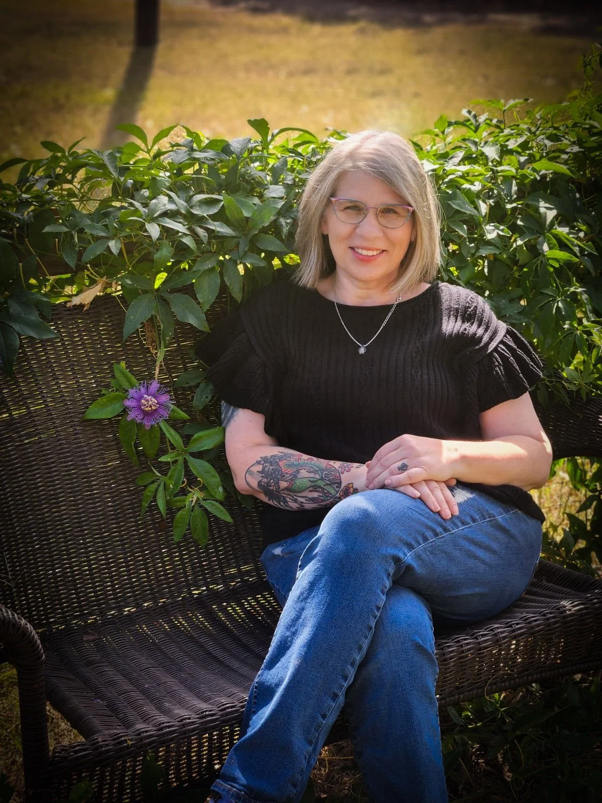 A woman with gray hair and glasses sitting on a wicker bench outdoors, smiling, with green plants and a pathway in the background.