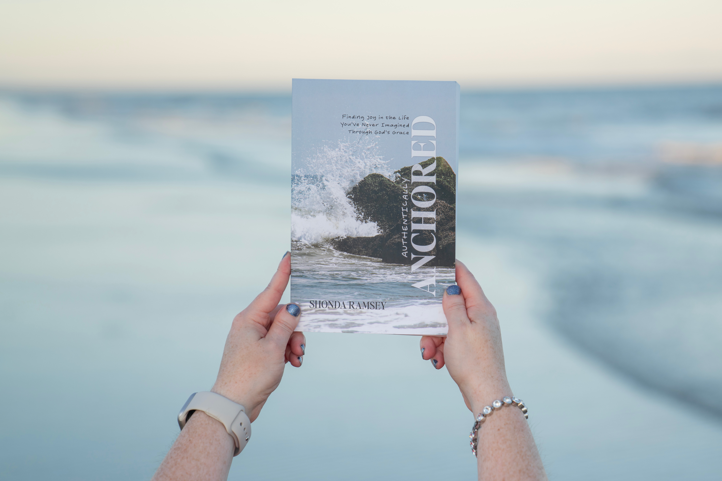Person holding a book titled "Anchored" by Shonda Ramsey, with a background of the ocean and waves.