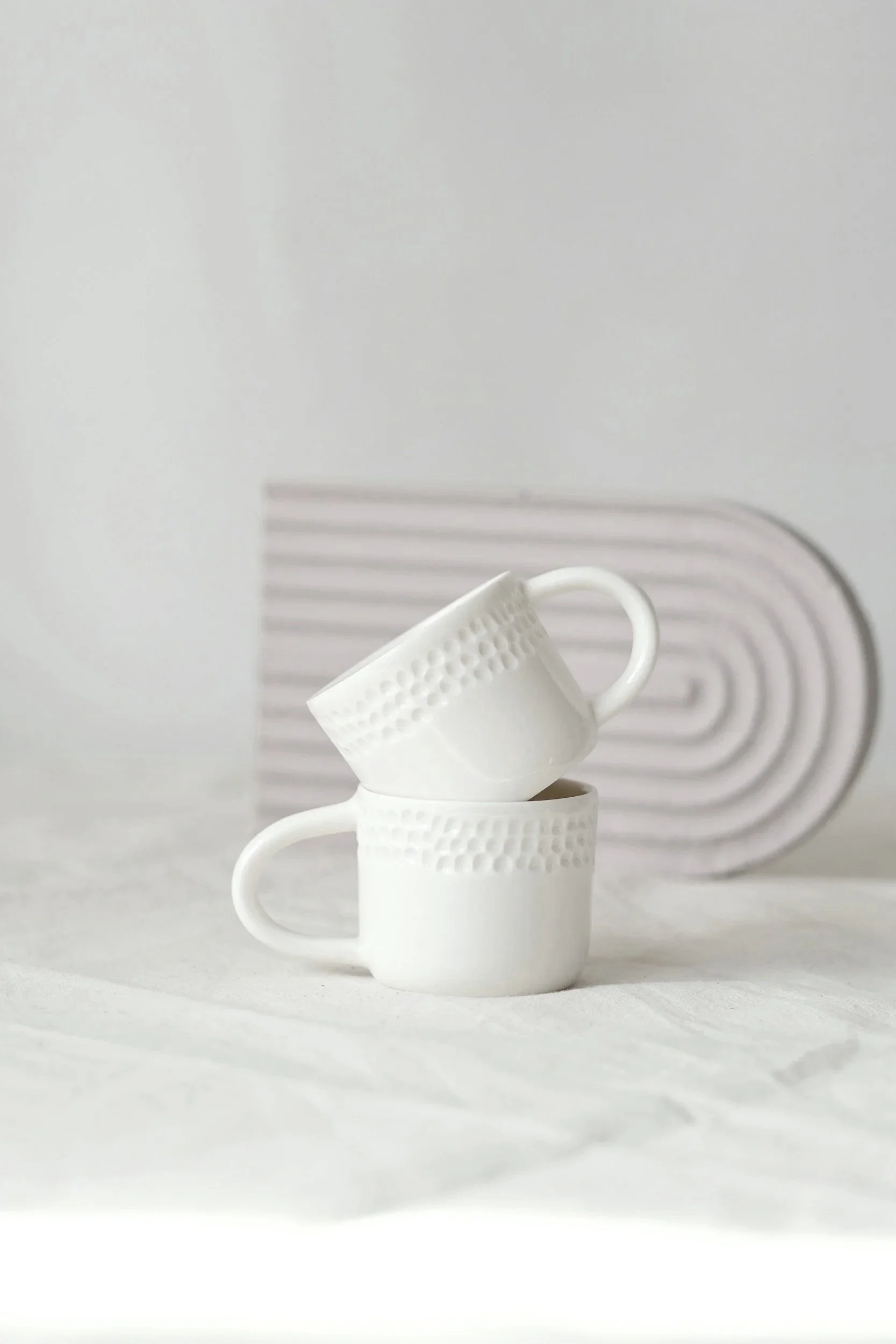 Three stacked white ceramic mugs with textured patterns and curved handles on a white surface against a light gray background.