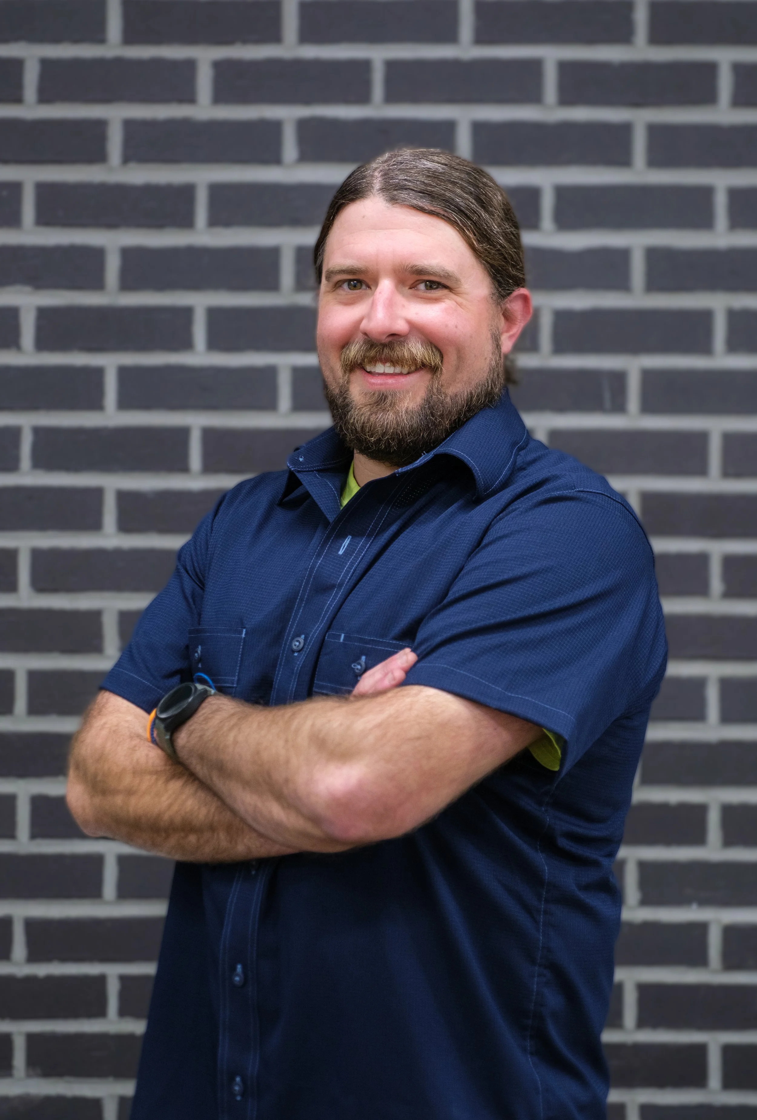 A man with long brown hair, beard, wearing a navy blue button-up shirt, standing with arms crossed in front of a black brick wall, smiling at the camera.