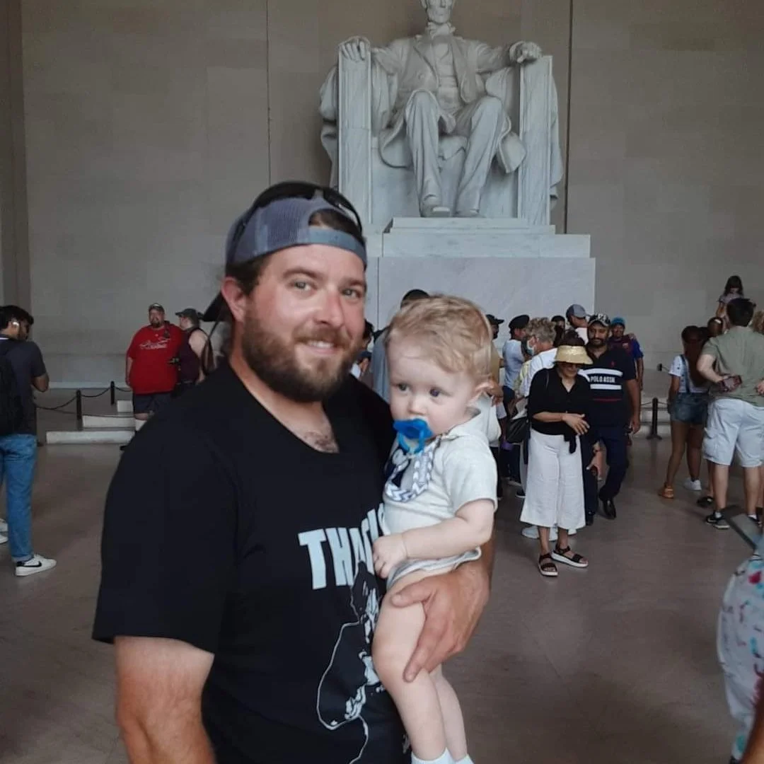 A man holding a young child with a pacifier in their mouth in the Lincoln Memorial in Washington, D.C. background includes tourists and the famous seated statue of Abraham Lincoln.