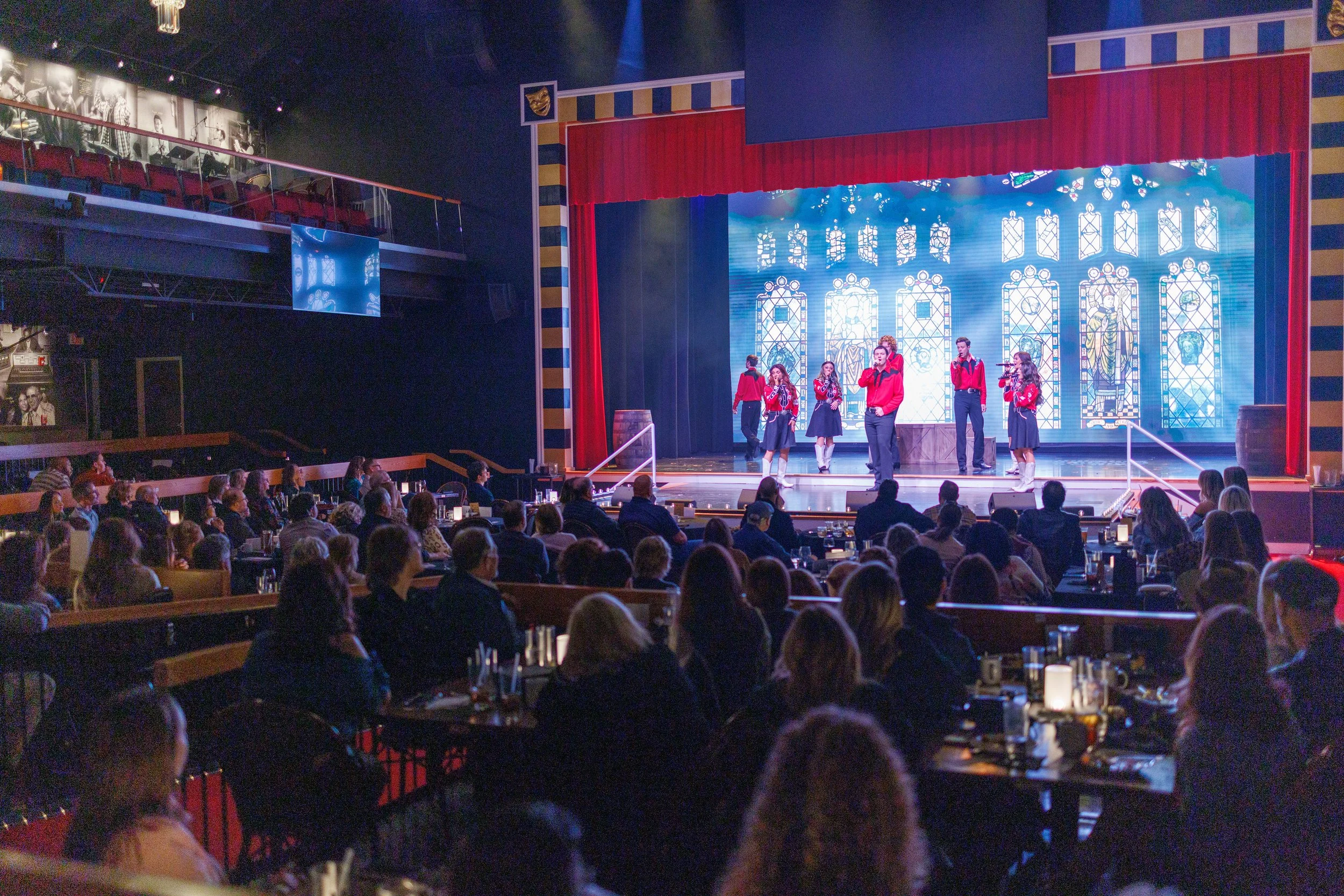 A stage with performers dressed in red and black costumes, singing into microphones, with a large stained glass window backdrop. The audience is seated at tables set with candles, in a theater or cabaret setting.