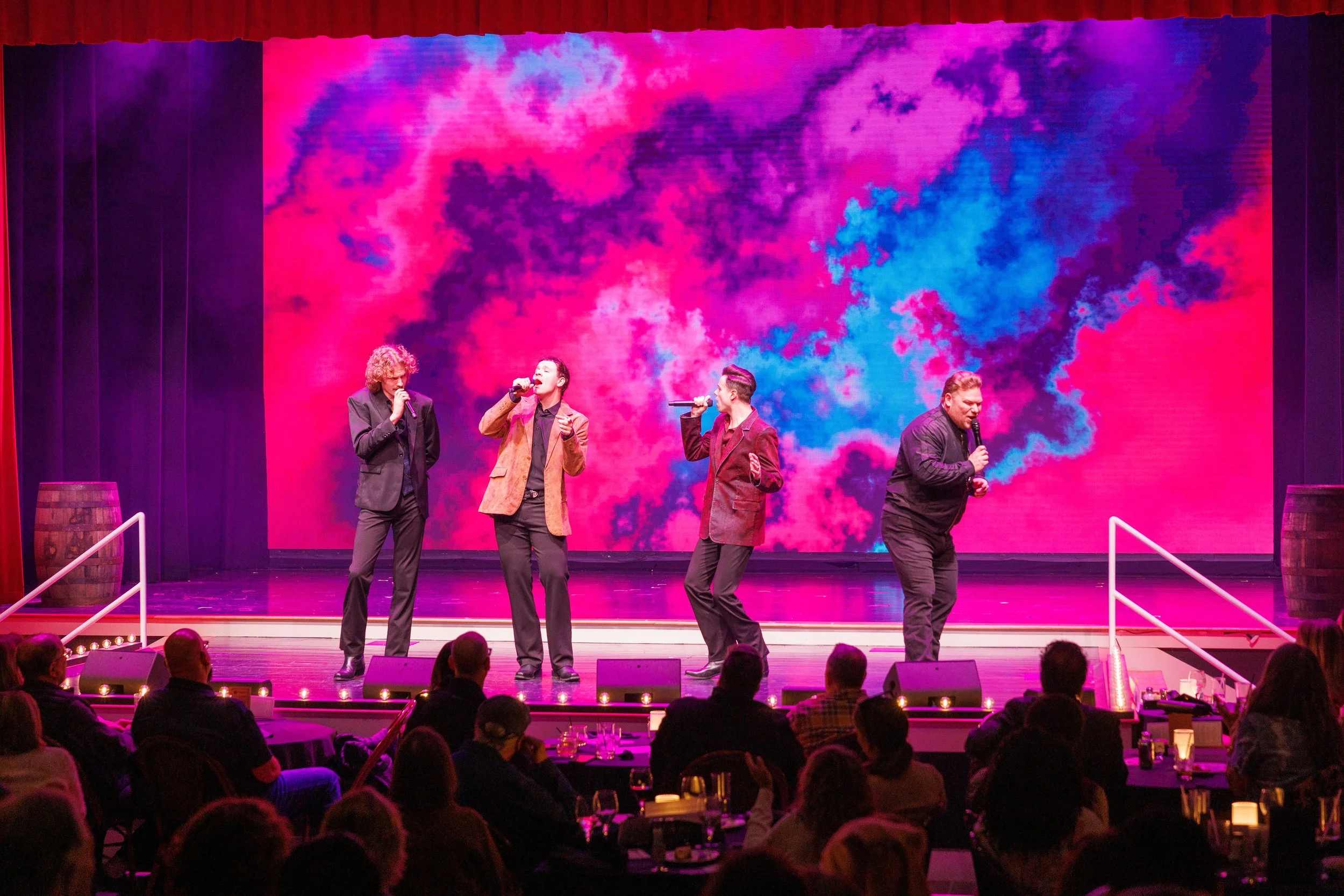 Four male performers singing on stage with a colorful digital background, audience watching, and stage props including barrels.