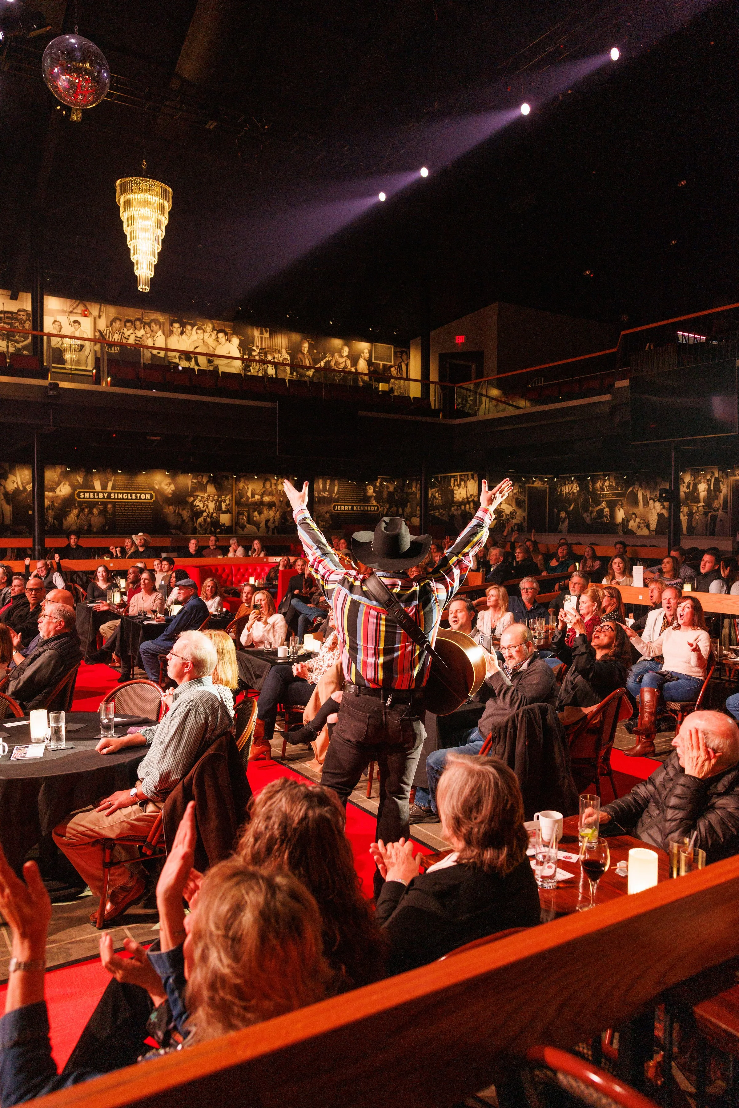 A performer on stage in a cowboy hat and colorful striped shirt, playing a guitar with arms outstretched, performing in front of a seated audience in a dimly lit venue with photographs on the walls and a large chandelier hanging from the ceiling.