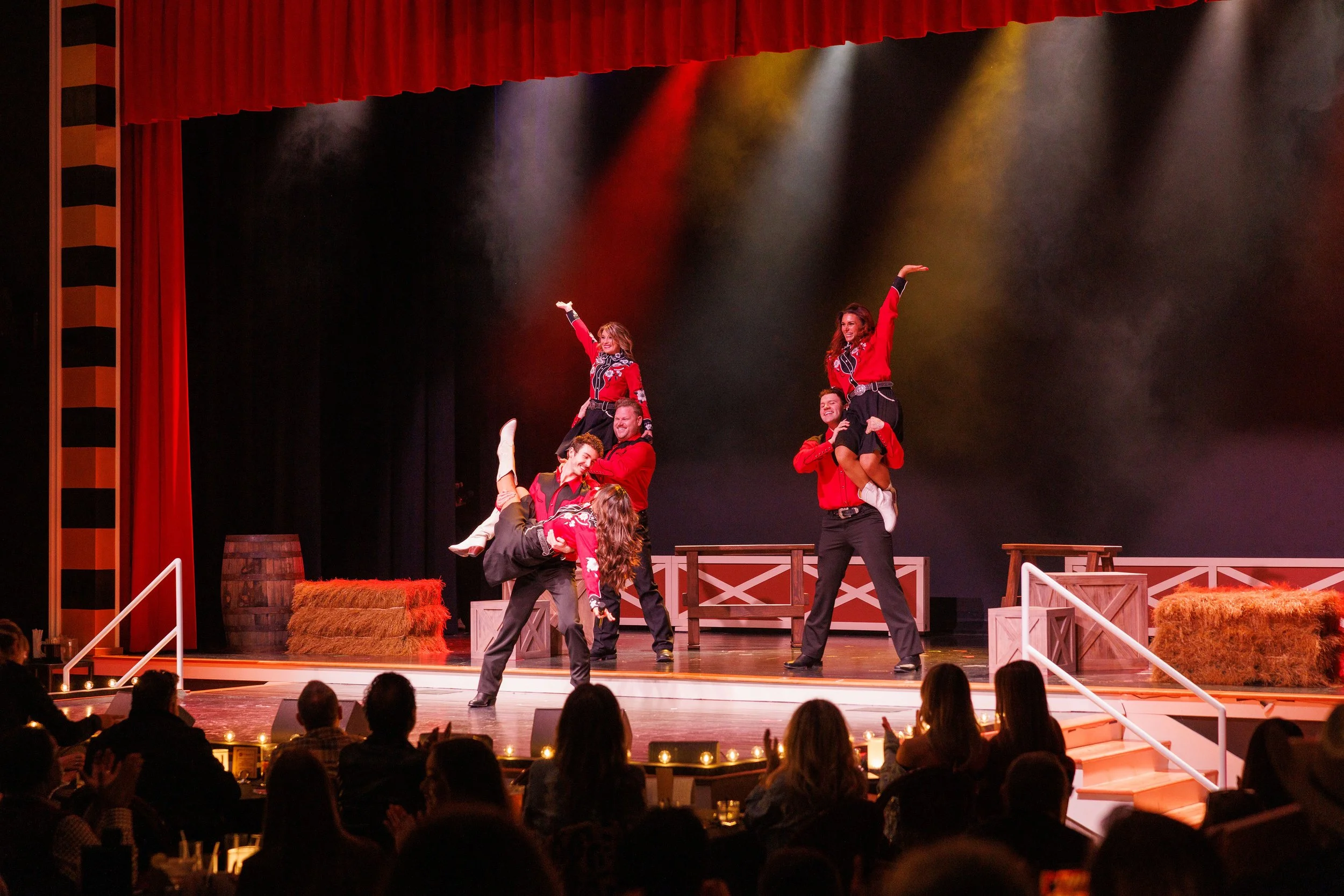 A group of performers dressed in Western-style outfits performing a choreographed dance on a theater stage. The stage has props like hay bales and wooden crates, with a red curtain above and an audience watching.