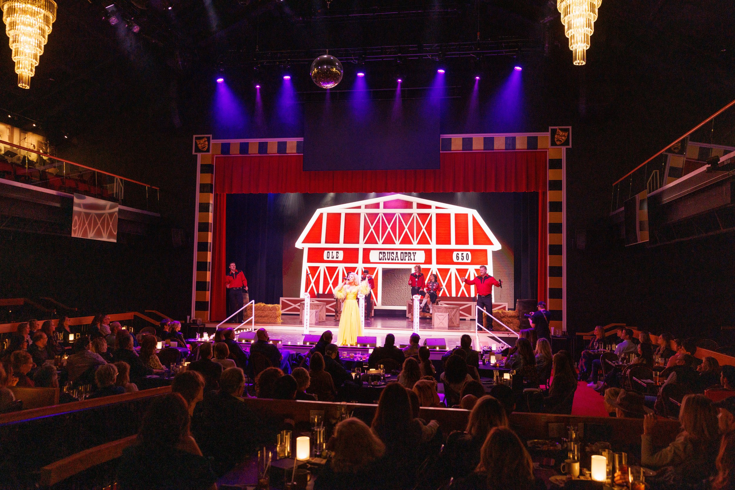A stage with a red barn-themed backdrop reading 'Ole Crusa Opry 650', surrounded by actors in cowboy costumes, under purple stage lights, with an audience seated at tables watching the performance.