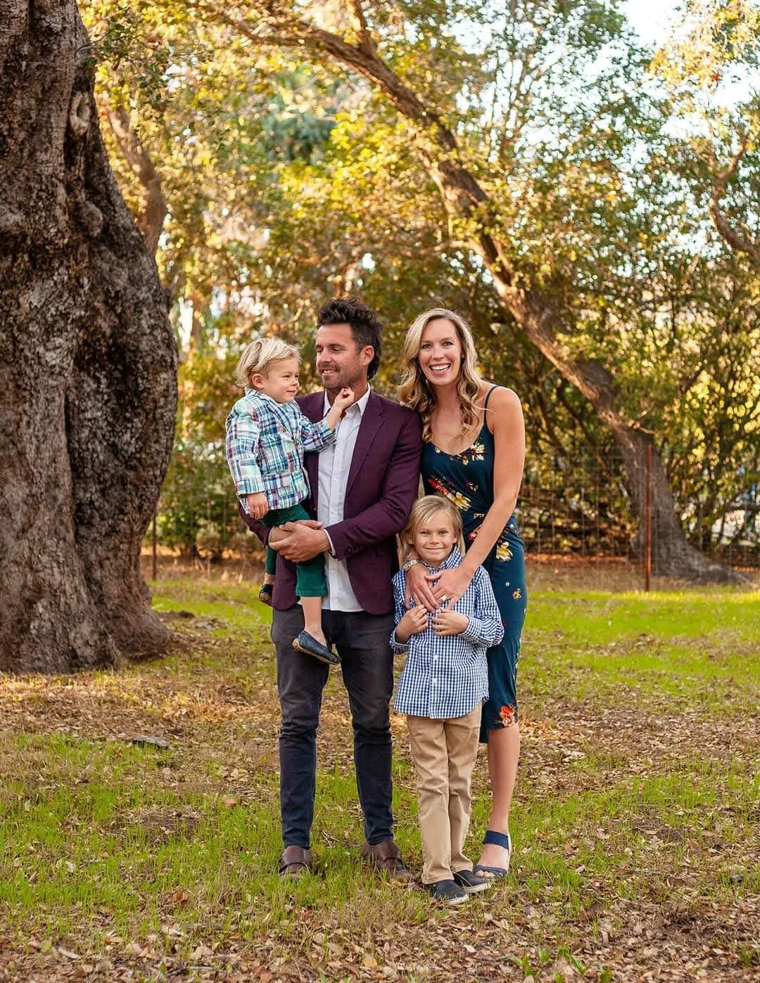 Picture of Family of Whitney Detar standing in a park with a large tree behind. One 4 year old blonde boy in a blue checkered suit jacket being held by dad in a maroon suit jacket. One blonde 9 year old blonde boy in a checkered blue shirt