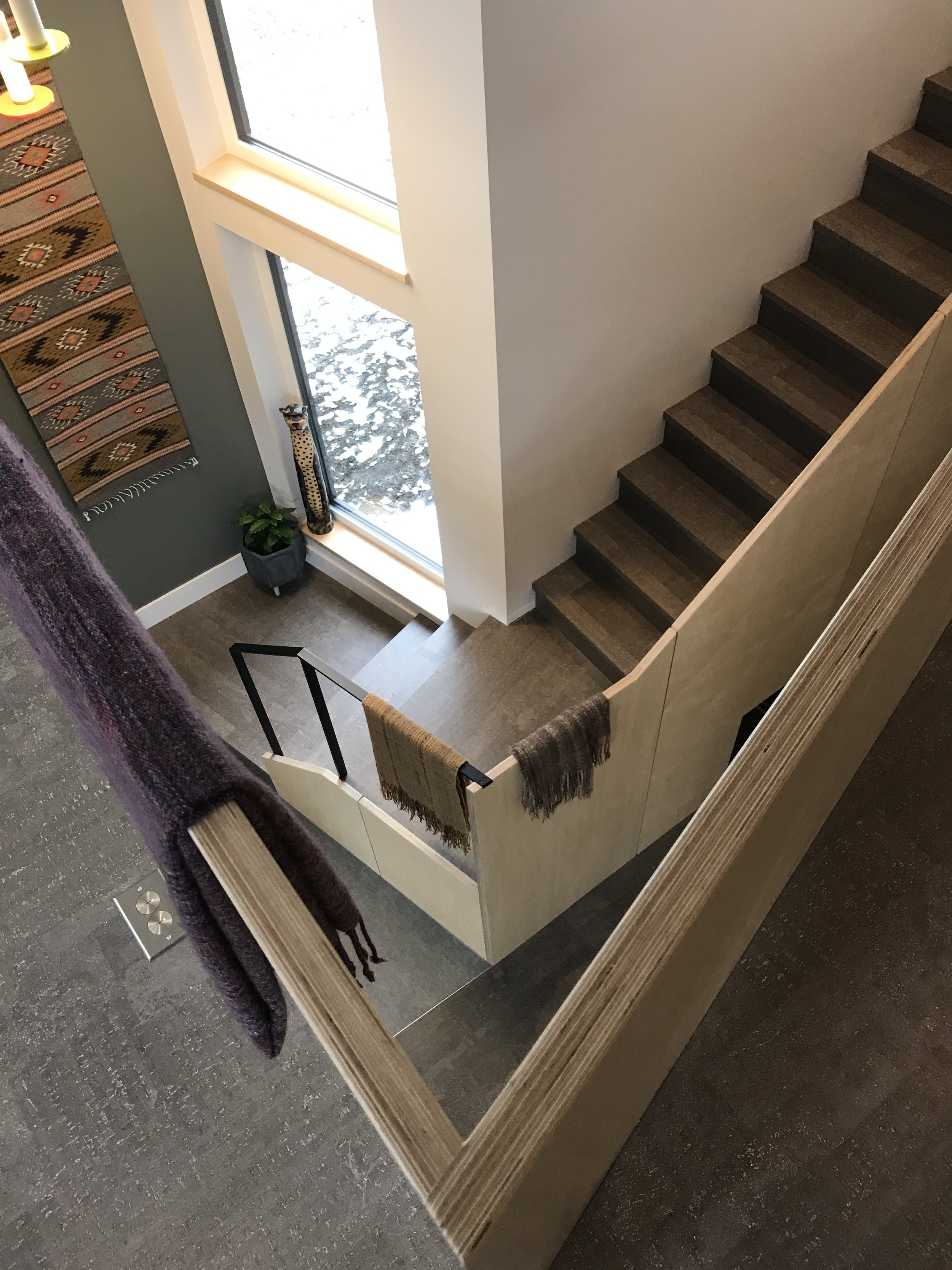 Modern interior with dark wood staircase, layered railing, woven textiles, snowy window view, and earthy-toned wall decor. Natural light and warm textures create a cozy, architectural ambiance.