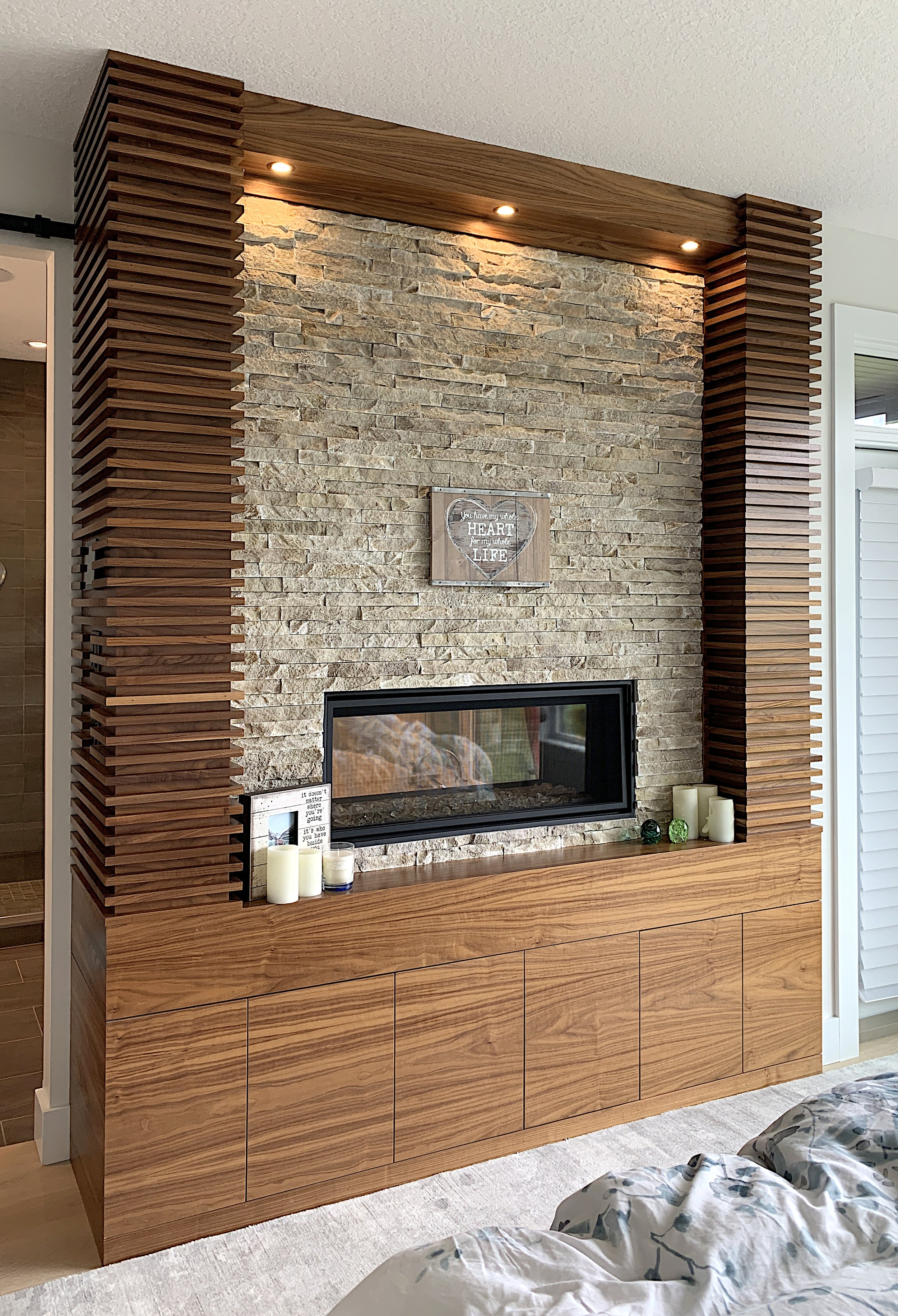 Modern bedroom fireplace with textured stone wall, vertical wood slats, recessed lighting, and decorative cabinet